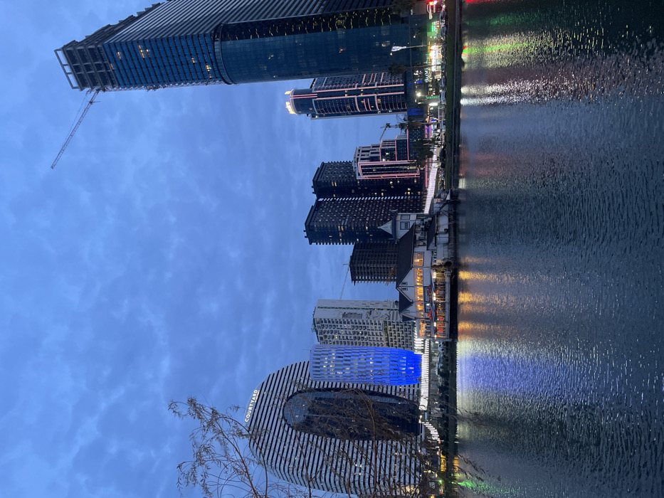 A waterfront cityscape with tall buildings reflecting in the water. Several illuminated skyscrapers line the shore, including one under construction with a visible crane. A building with a prominent "COURTYARD" sign is visible in the foreground. The water mirrors the lights, creating streaks of color. The sky is a dusky blue, indicating either twilight or early evening.