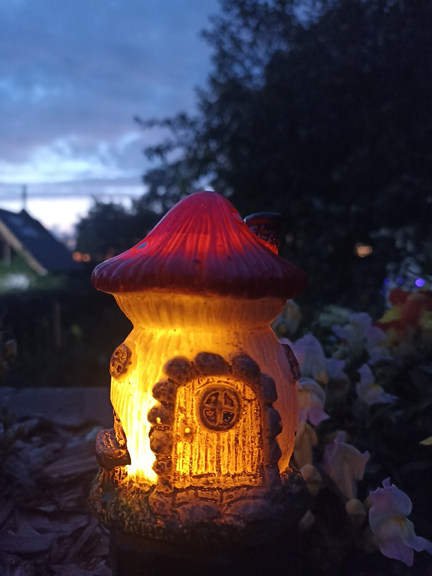 A close-up, outdoor, eye-level shot captures a lit, mushroom-shaped garden ornament at dusk. The ornament is primarily yellow with a red cap and features a detailed, small door and windows. Behind the ornament, blurry elements suggest a garden with some flowering plants, trees, and a house with a dark roof. The sky above is a deep blue, indicating twilight. The image is set in the city of Mochische, Russia.