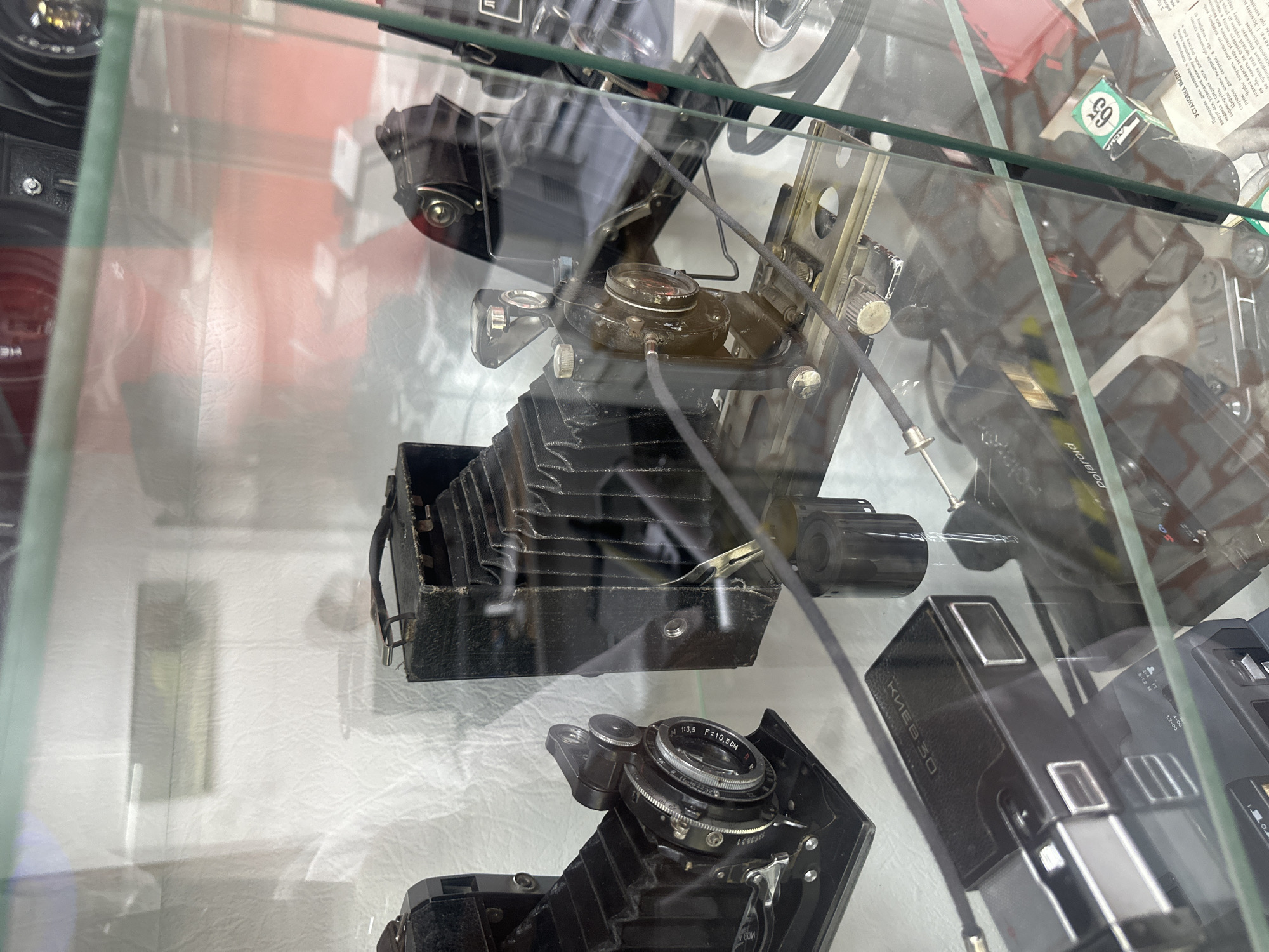 This image depicts an indoor display case filled with vintage cameras. The display appears to be located in the Tobolsk Museum of Local Lore (Tobolskiy muzey-zapovednik), a cultural institution in Tobolsk, Russia. The cameras are varied in design, with several notable examples including a black bellows camera on the left, another larger black bellows camera to its right, and a smaller, more modern-looking black camera with silver accents at the bottom left. A roll of film is visible next to the larger bellows camera. The cameras are arranged on shelves within a glass-fronted display, suggesting a museum or antique shop setting. Reflections on the glass suggest an overhead lighting source and possibly other exhibits in the vicinity. The background is a textured white surface, possibly a backdrop or wall. There are no people visible in the image.