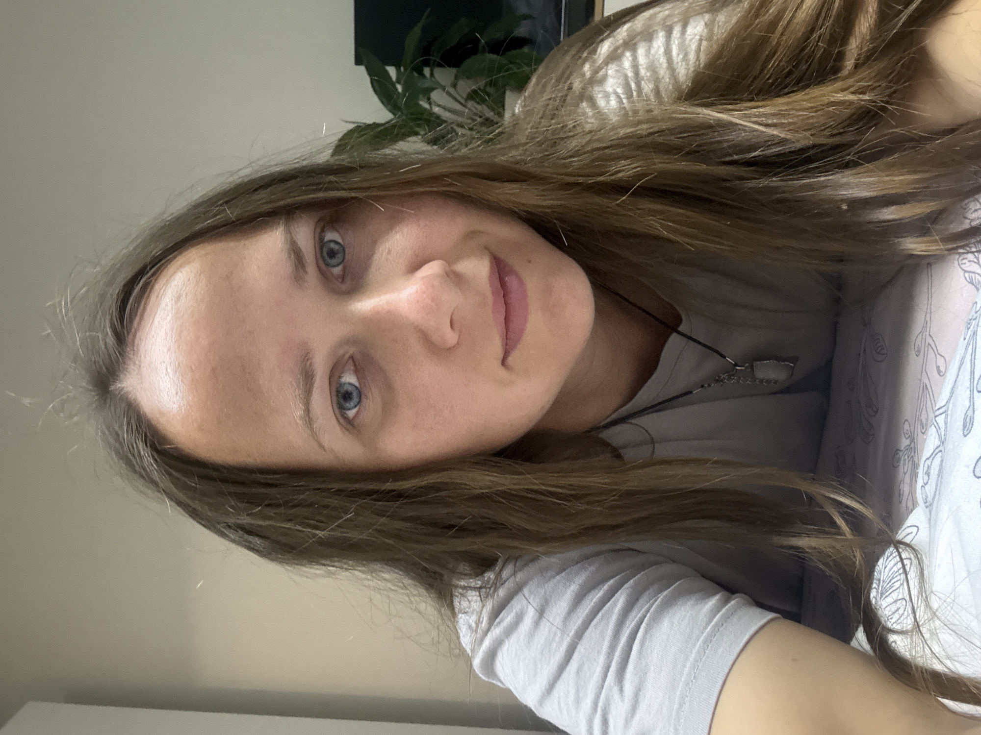 A head-and-shoulders, front-facing selfie shows a woman with long, light brown hair, blue eyes, and fair skin. She is wearing a gray t-shirt and a necklace with a light-colored pendant. The background is out of focus and appears to be an indoor setting, possibly a bedroom or living room, with a wall and a plant visible. The lighting suggests it is daytime. The image was taken in Izhevsk, Russia.