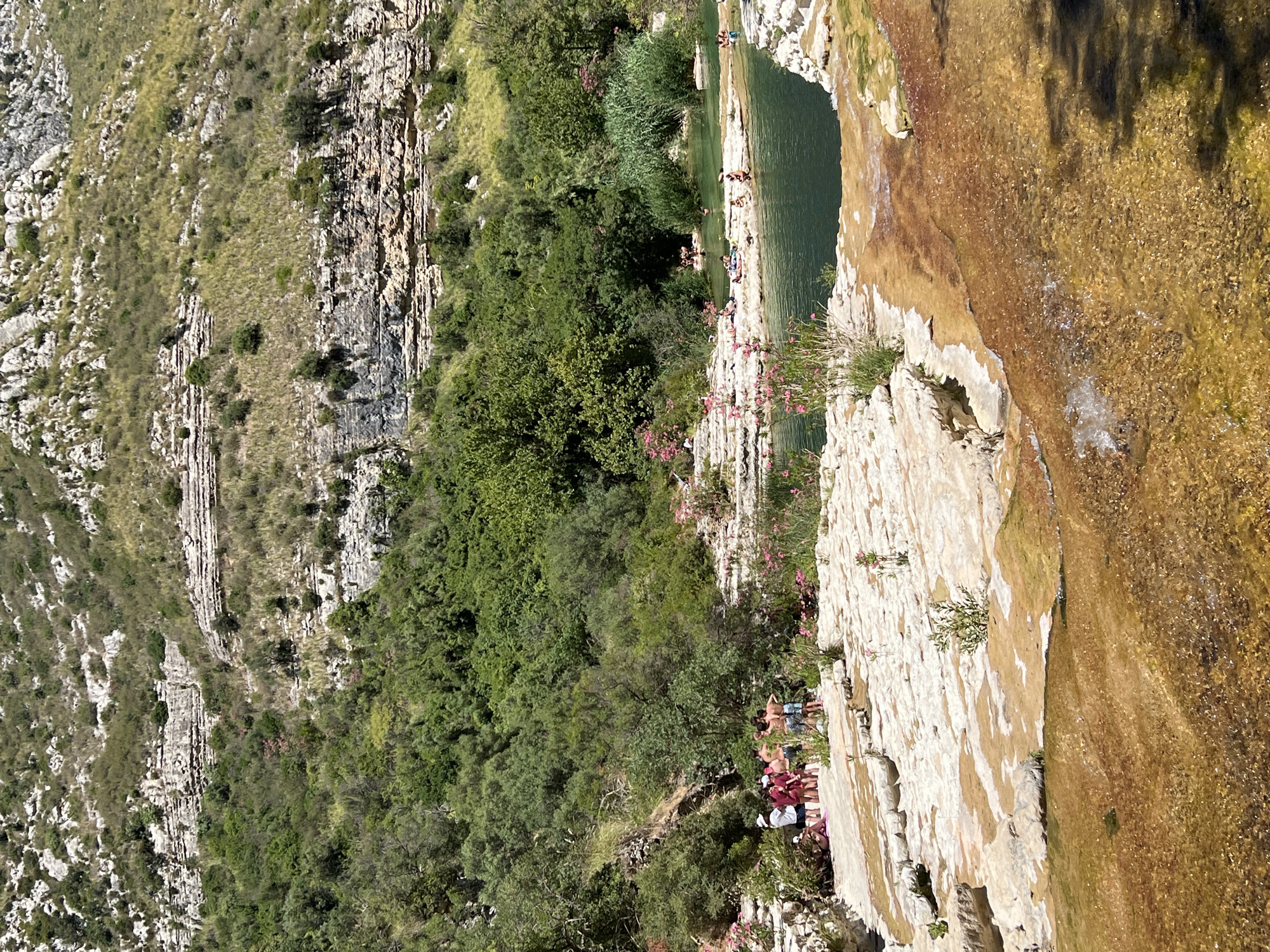 The image depicts a natural landscape featuring a river with clear, greenish-blue water flowing over textured, light-colored rocks. Several people are visible relaxing and swimming in the river and on the surrounding rocky terrain. Lush green trees and vegetation cover the hillsides, with rocky formations and striations visible on the slopes in the background. The scene appears to be a popular outdoor recreational spot, possibly within the city of Noto, Italy, known for its natural beauty. The water is shallow in some areas, revealing the rocky riverbed, while other parts are deeper and more shaded by the surrounding foliage. The overall impression is one of a serene and scenic natural environment with human activity integrated into the landscape.