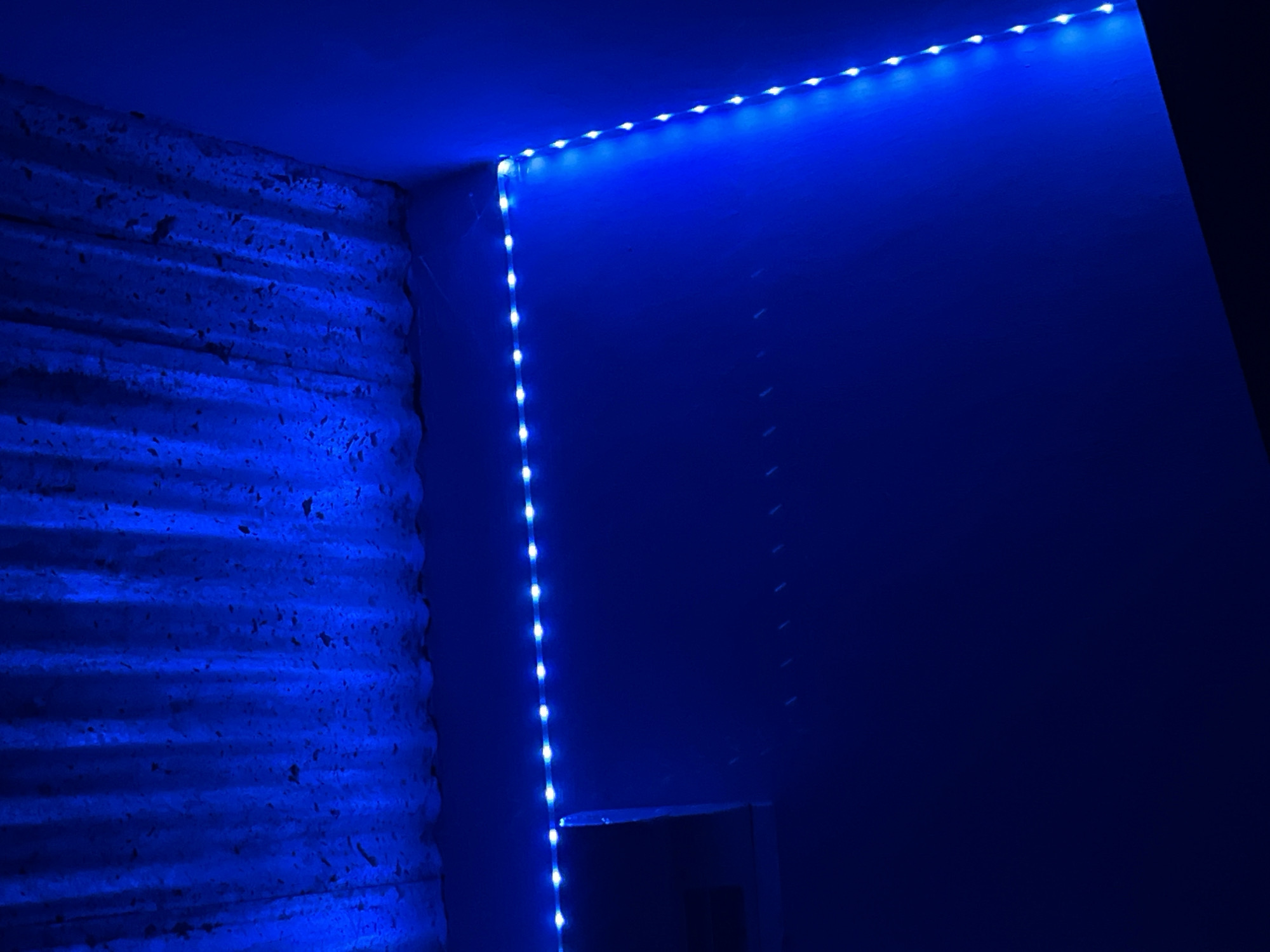 The image shows a dark room illuminated by blue LED strip lights. The lights are mounted along the ceiling and down a wall in the corner. The ceiling appears to be made of corrugated metal. There is a dark rectangular object mounted on the wall near the ceiling on the left side of the image. The room is otherwise featureless and dark. No people or animals are visible. This scene is likely from a residential or storage space in Juan Griego, Venezuela, possibly a home or an industrial area, given the corrugated metal ceiling.