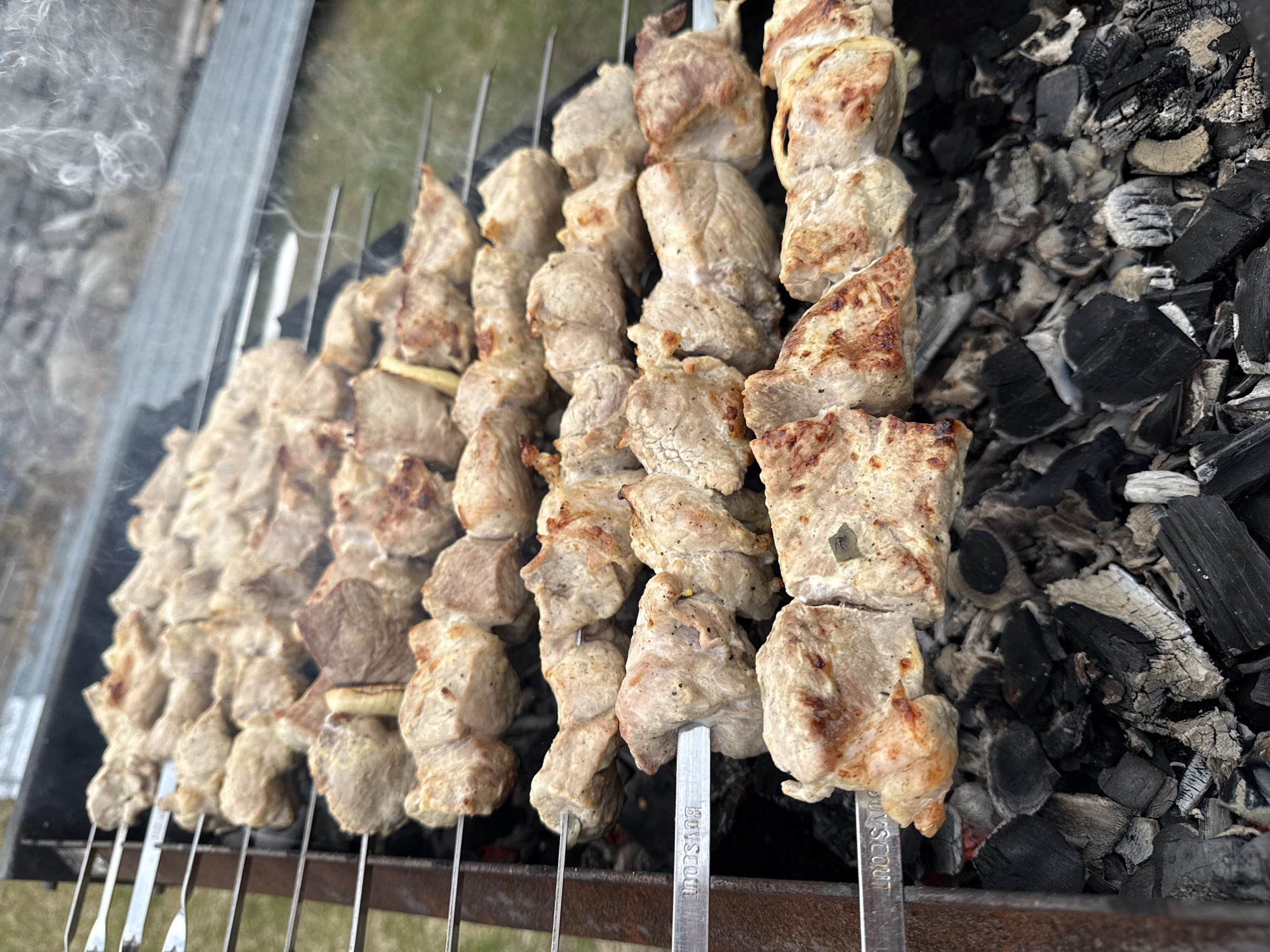 The image shows meat skewers cooking on a grill in what appears to be an outdoor setting. The location is Верхнеказымский, Russia. The skewers are metal and hold chunks of meat that are in varying stages of being cooked, ranging from lightly browned to more thoroughly cooked. The grill contains hot coals that are actively burning, providing the heat for cooking. The skewers are arranged in a row over the coals, suggesting they are being cooked simultaneously. The scene appears to be a backyard or park setting, with a glimpse of grass and trees in the background.
