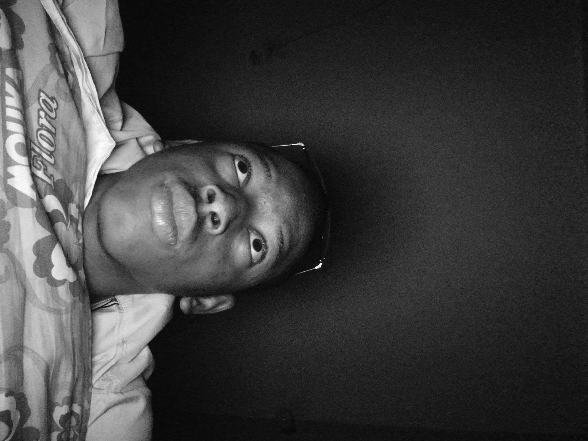 A low-angle, black-and-white, close-up photograph shows a young Black male looking upwards, with his head tilted back. He is wearing glasses perched on his head and a light-colored hooded jacket over a patterned shirt. The background is dark and indistinct, suggesting an indoor setting with minimal light. The focus is on his face, particularly his eyes and lips, with the rest of his body partially obscured by the bedding in the foreground. The bedding has a floral pattern with text that appears to read "FLORA MOLINA".