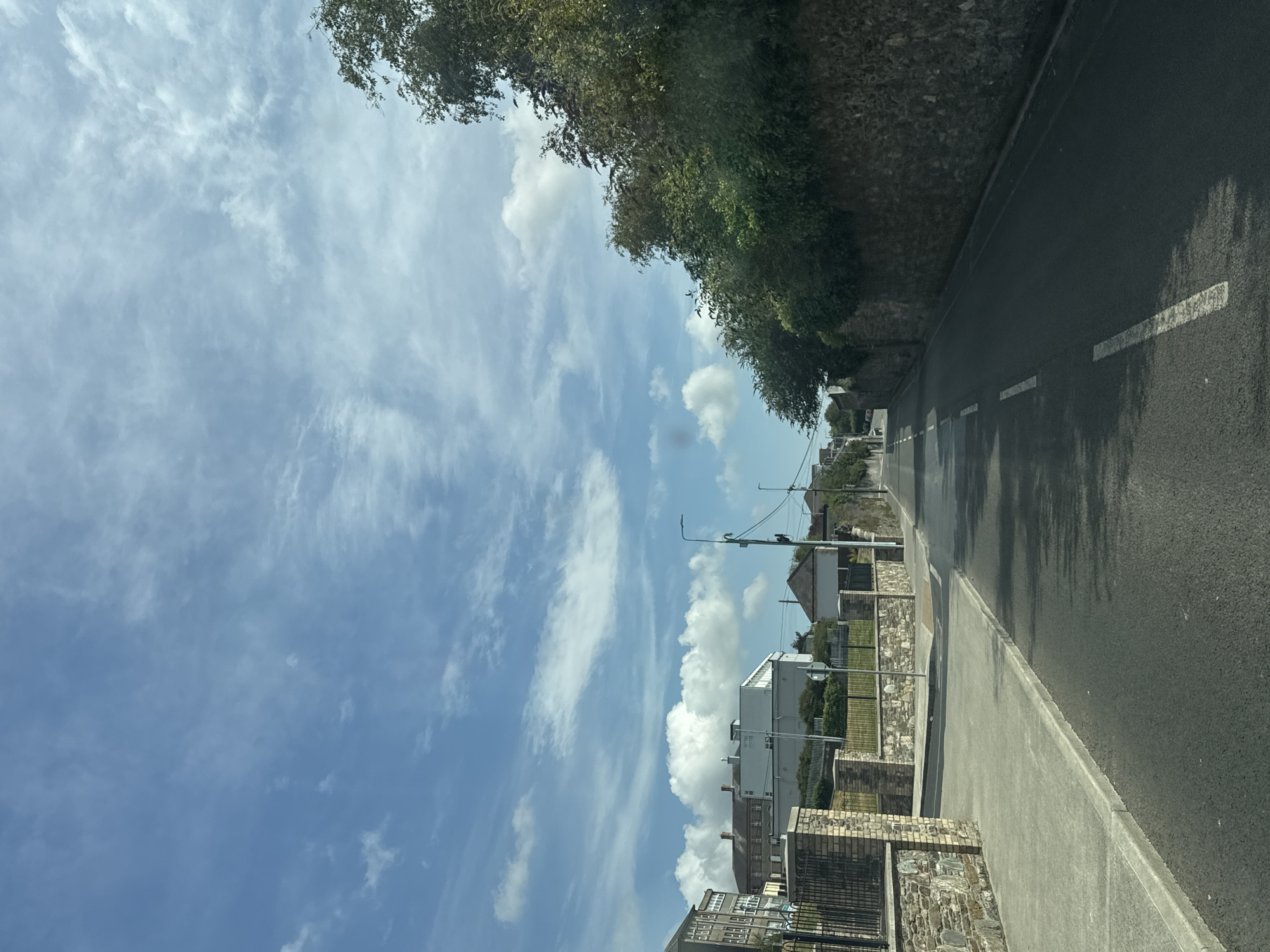 This image captures a sunlit street scene, likely in the city of Waterford, Ireland. The perspective is from a vehicle driving along a paved road with white dashed lane markings. On the left side of the road, a concrete sidewalk runs alongside a stone wall and a black metal fence. Beyond the fence, there are several buildings visible, including a multi-story brick structure and some white modern buildings.
To the right of the road, a thick hedge of green trees grows atop a weathered stone wall, which extends into the distance. The sky above is a vibrant blue, dotted with wispy white clouds and some larger, fluffier cumulus formations. The overall impression is of a quiet residential or commercial street on a pleasant day. The presence of streetlights and the general architecture are consistent with urban or suburban areas in Ireland.
