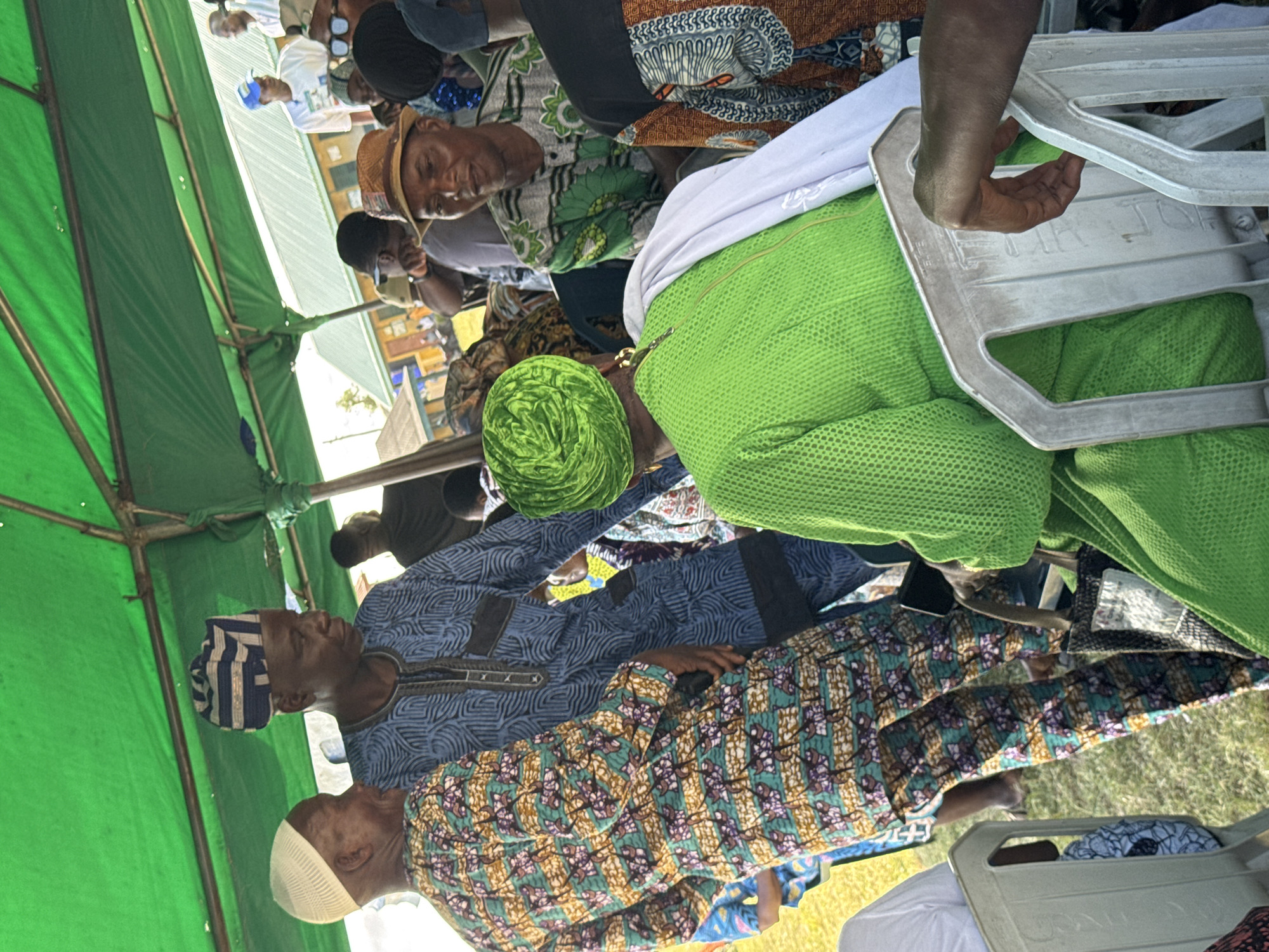 A gathering is taking place in what appears to be Badagry, Nigeria, based on the style of dress and headwear of the individuals present. Several people are seated under a green tent structure, possibly for an event or ceremony. The individuals are wearing patterned and colored clothing, with some wearing traditional Yoruba hats (fila). The scene features various chairs, and there is grass visible in the background. The focus of the image is on the people and their clothing.