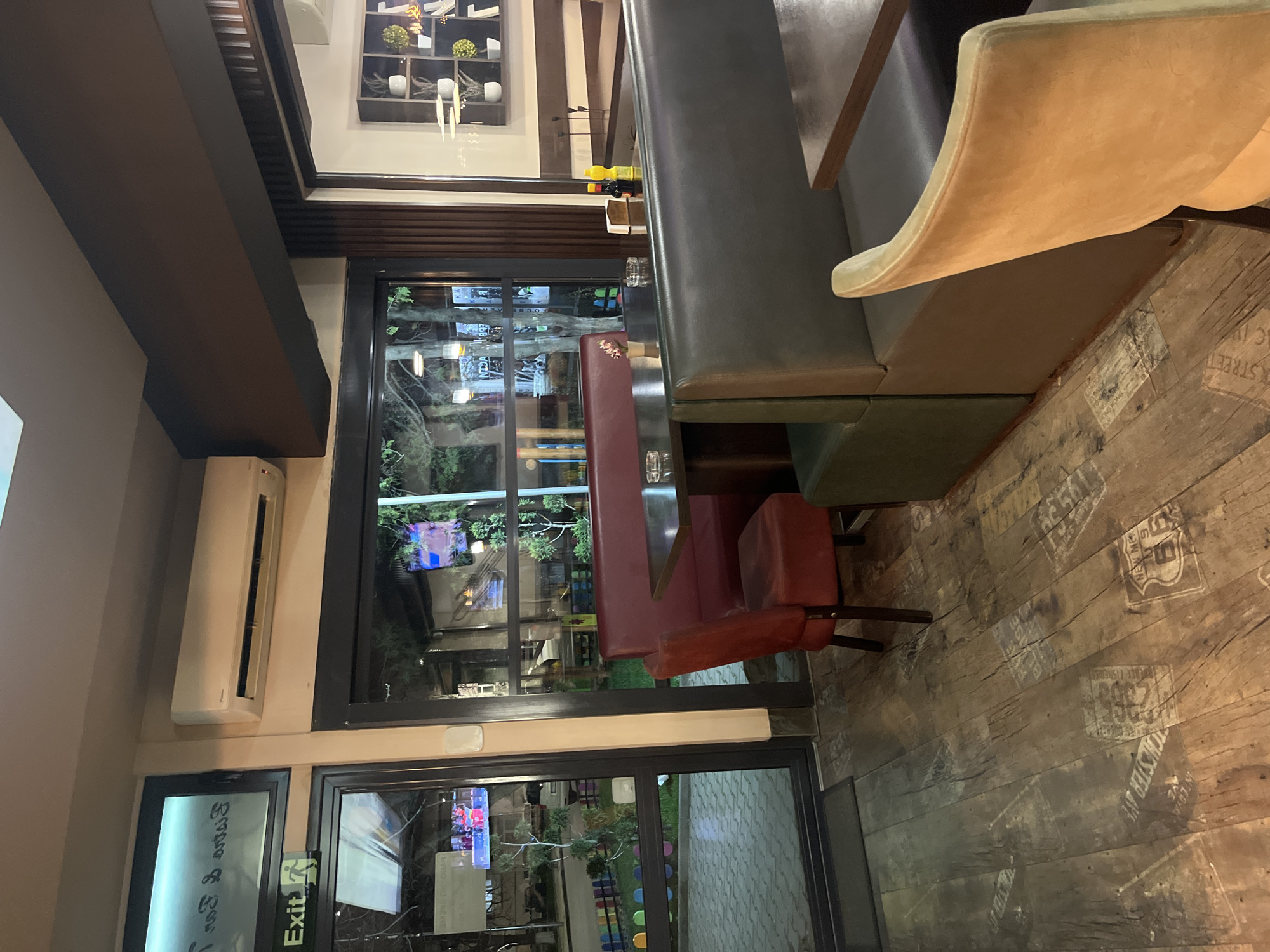 A restaurant interior in Nova Zagora, Bulgaria, features dark wood plank flooring with faded stenciled street names. Booth seating with a high gray back and green base sits to the right of a red booth with clear acrylic accents. A tan-colored chair is partially visible. Large windows offer a view outside, with reflections of trees and buildings. An air conditioning unit is mounted on the wall near an exit sign. The lighting is a mix of natural light from the windows and artificial light, creating a bright interior.