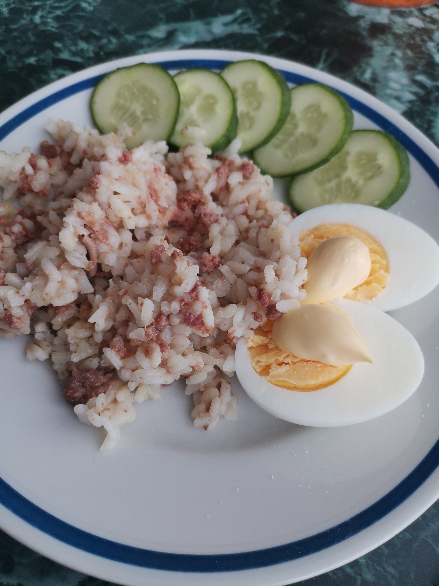 A plate of food sits on a table with a dark green, marbled surface. The plate contains a serving of white rice mixed with small pieces of reddish-brown meat, sliced cucumber rounds, and two halves of a hard-boiled egg topped with mayonnaise. The plate has a blue band around its rim.