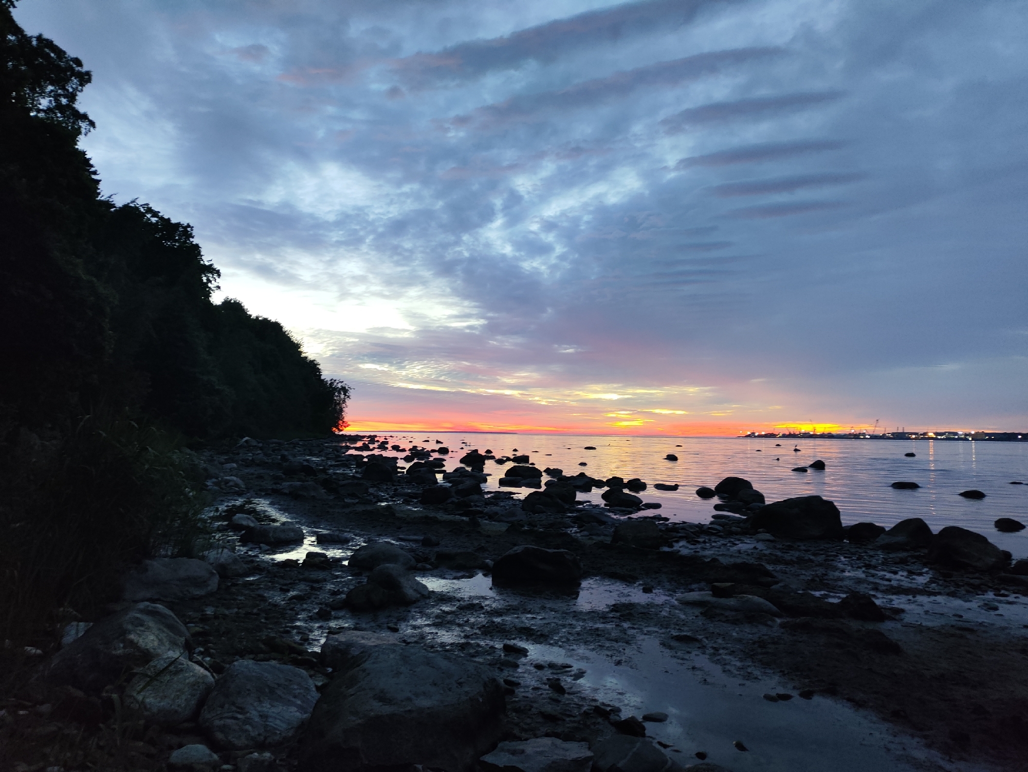 A serene sunset over the Baltic Sea, as seen from a rocky shoreline in Tallinn, Estonia. The sky is painted with hues of orange, pink, and purple, with dramatic clouds stretching across the horizon. The water reflects the colorful sky, creating a mirror-like effect. In the foreground, dark, wet rocks and patches of mud are visible, with a few scattered boulders. A dense line of dark trees hugs the coastline on the left side of the image. In the distance, a faint outline of a city skyline, likely Tallinn's port area, with industrial structures and lights, can be observed. The overall atmosphere is peaceful and tranquil, capturing the end of a day in a coastal city.