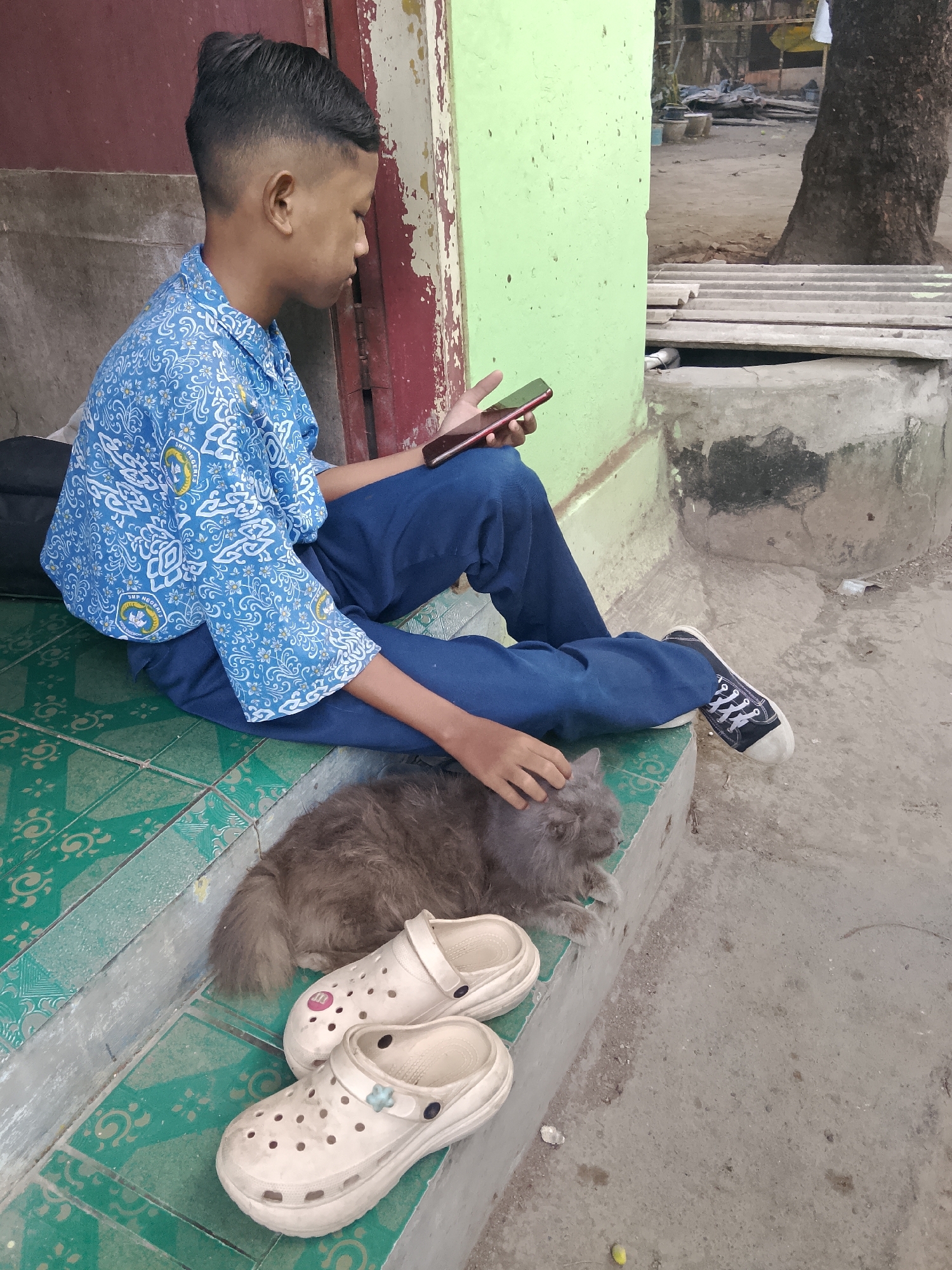 A young boy in a blue school uniform with a white patterned design sits on green tiled steps, looking at his phone. He is wearing dark blue pants and black and white sneakers. A gray, long-haired cat lies on the steps next to him, and he is petting it with his right hand. In front of the cat are two white clogs. The scene appears to be outdoors, with a green wall to the boy's left and a concrete ground and a tree to the right. The boy's uniform has a school emblem that reads "SMP NEGERI 1 CIREBON," indicating the location is Cirebon, Indonesia.