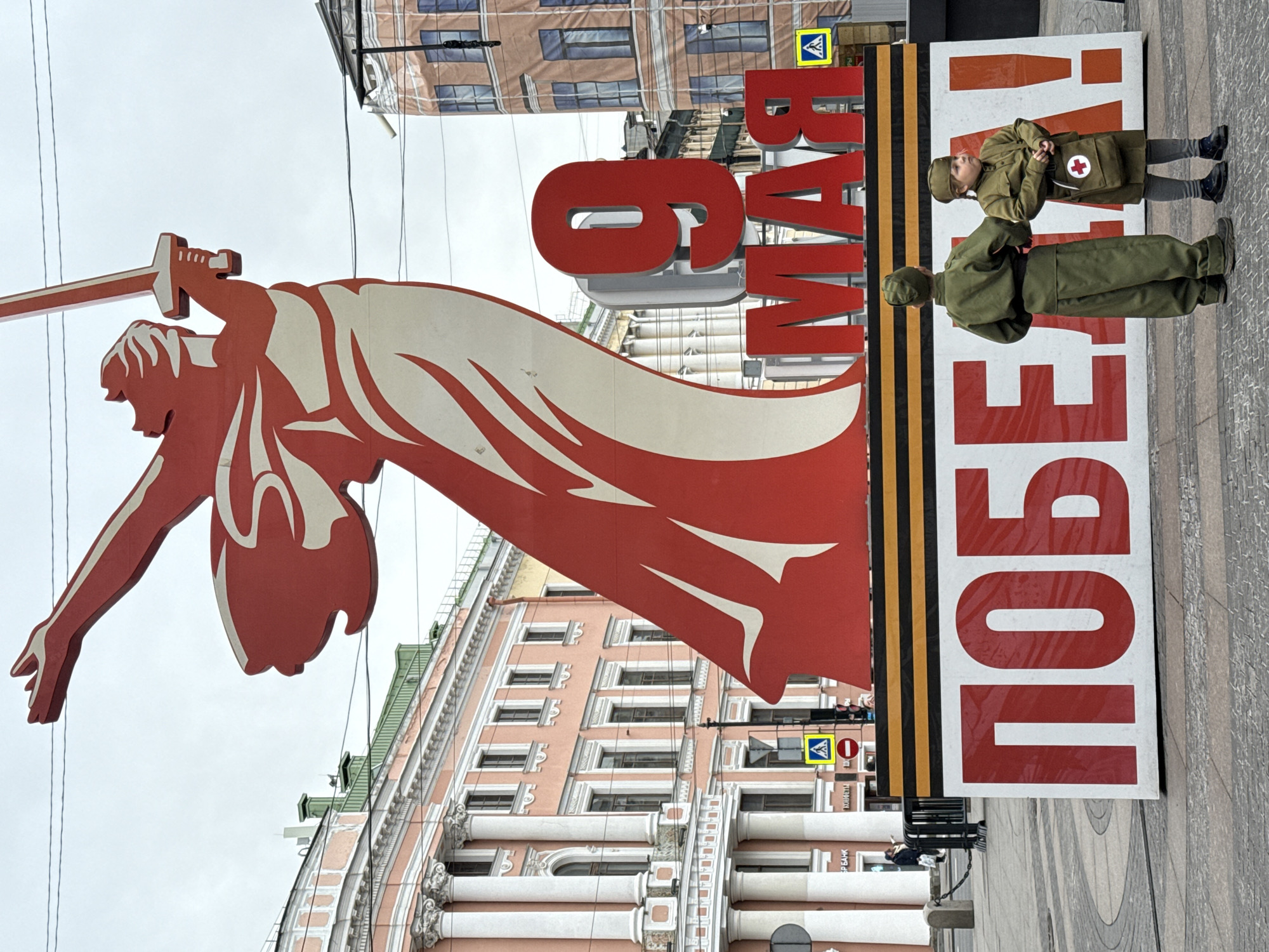 Here is a description of the image:
The image is taken in Saint Petersburg, Russia and appears to be celebrating Victory Day. A large, stylized, red cutout of the Motherland Calls statue holding a sword is prominent. Next to it is a sign with a Saint George ribbon and Russian text "9 MAЯ ПОБЕДА", which translates to "May 9 Victory". Two individuals in green military-style uniforms are posing near the sign. A girl is dressed as a medic, with a red cross on her bag. The background shows buildings with classical architectural features, and pedestrian crossing signs.