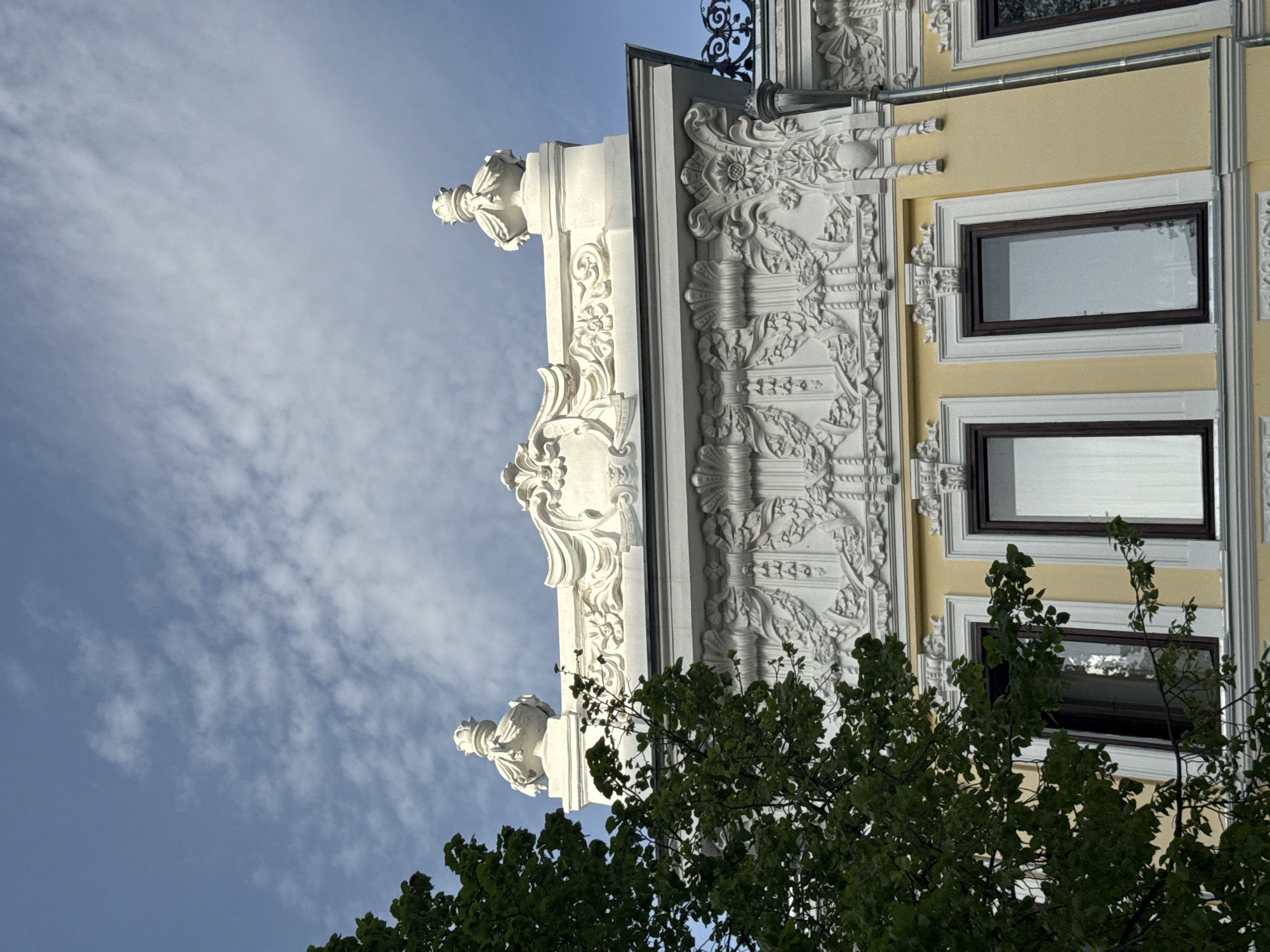 The image shows a section of a building facade in Kazan, Russia. The building features intricate white ornamental details, including decorative molding, sculpted foliage and finials. The facade has yellow painted sections surrounding rectangular windows with white frames and dark window borders. The sky is light blue with scattered white clouds in the upper left portion. A green tree with dense foliage obscures part of the lower right and bottom of the building.