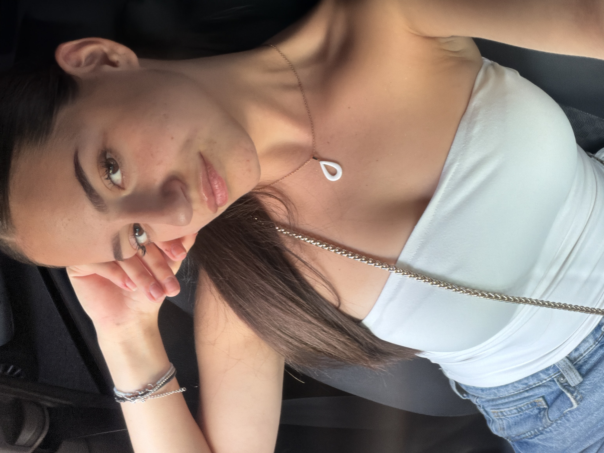 A young woman with long, dark brown hair is taking a selfie in what appears to be the interior of a car, likely in Burgas, Bulgaria, given the prompt's context. She is wearing a white strapless top and blue denim jeans. She has a delicate necklace with a white teardrop pendant around her neck and a bracelet on her left wrist. Her left arm is propped up, with her hand resting on her forehead, as she looks towards the camera. The background is dark and indistinct, suggesting the car's interior.