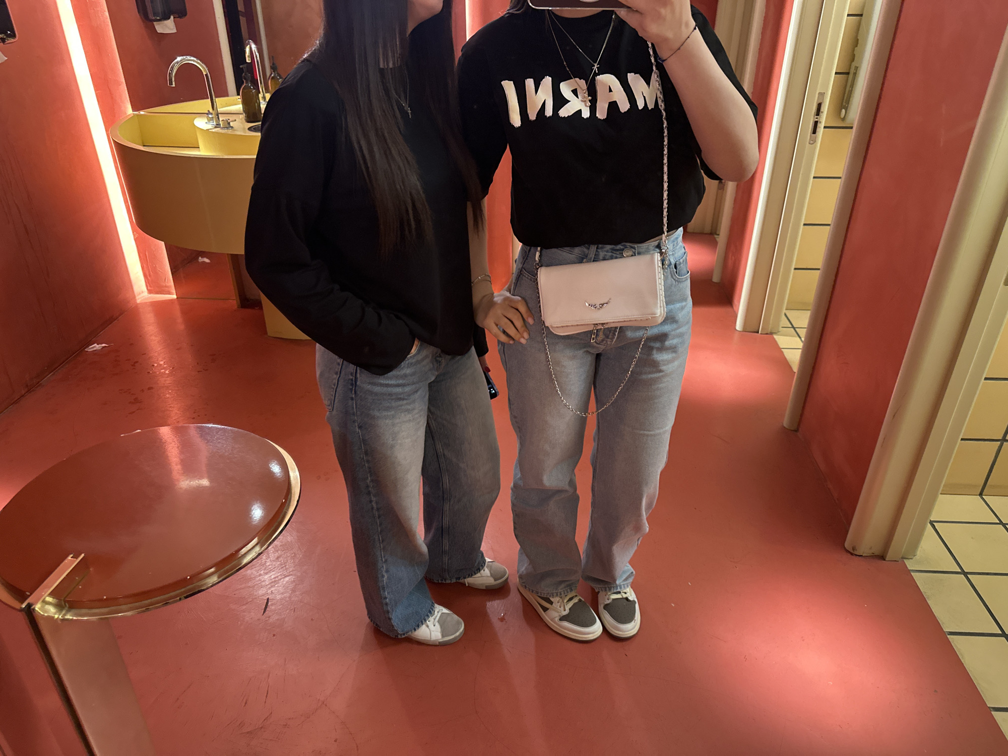 Two young women stand side-by-side in what appears to be a public restroom. The woman on the left wears a long-sleeved black shirt and light blue jeans with grey and white shoes. The woman on the right wears a black t-shirt with white letters, light blue jeans, and grey and white shoes. She also has a small white purse with a silver chain. The restroom has a red floor and walls, a yellow sink, and a round, brown, table-like object. There are doorways in the background with tan and white trim. This location cannot be independently verified as being in Yerevan, Armenia based on the given image.