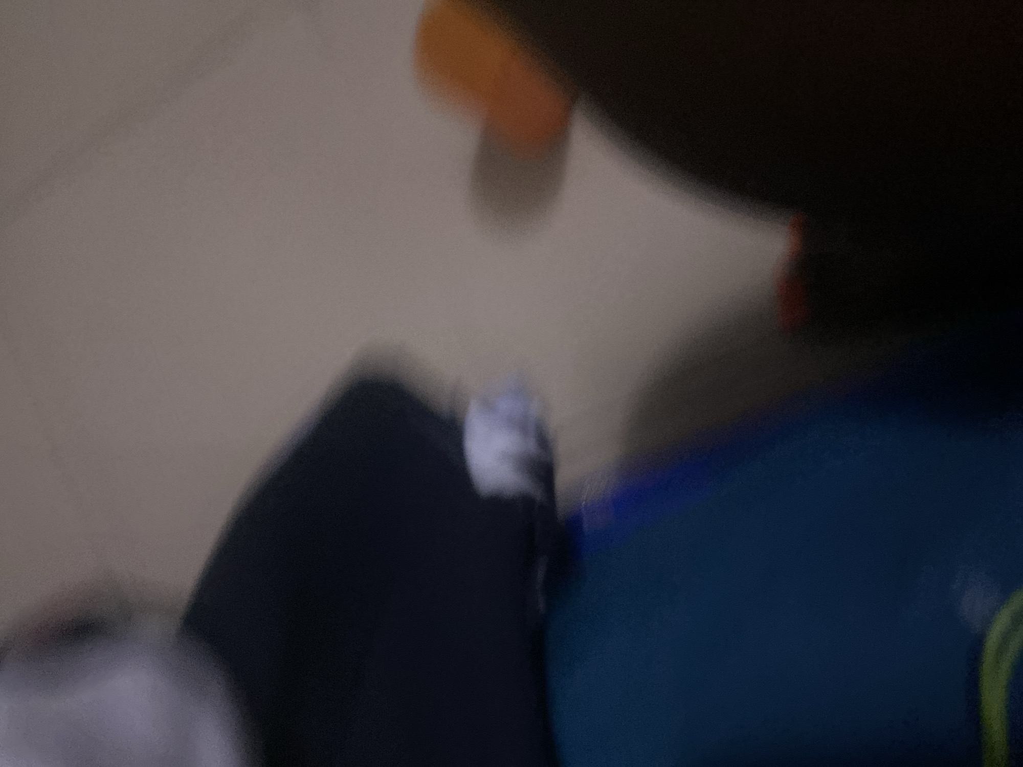 In a dimly lit indoor setting, an out-of-focus individual wearing a dark shirt and light-colored cuffs stands beside a large, dark blue object. To the right, a blurred orange object is visible, casting a shadow on the wall behind. The background appears to be a neutral-colored wall. No specific location in Howayi, Nigeria can be determined from the provided image.