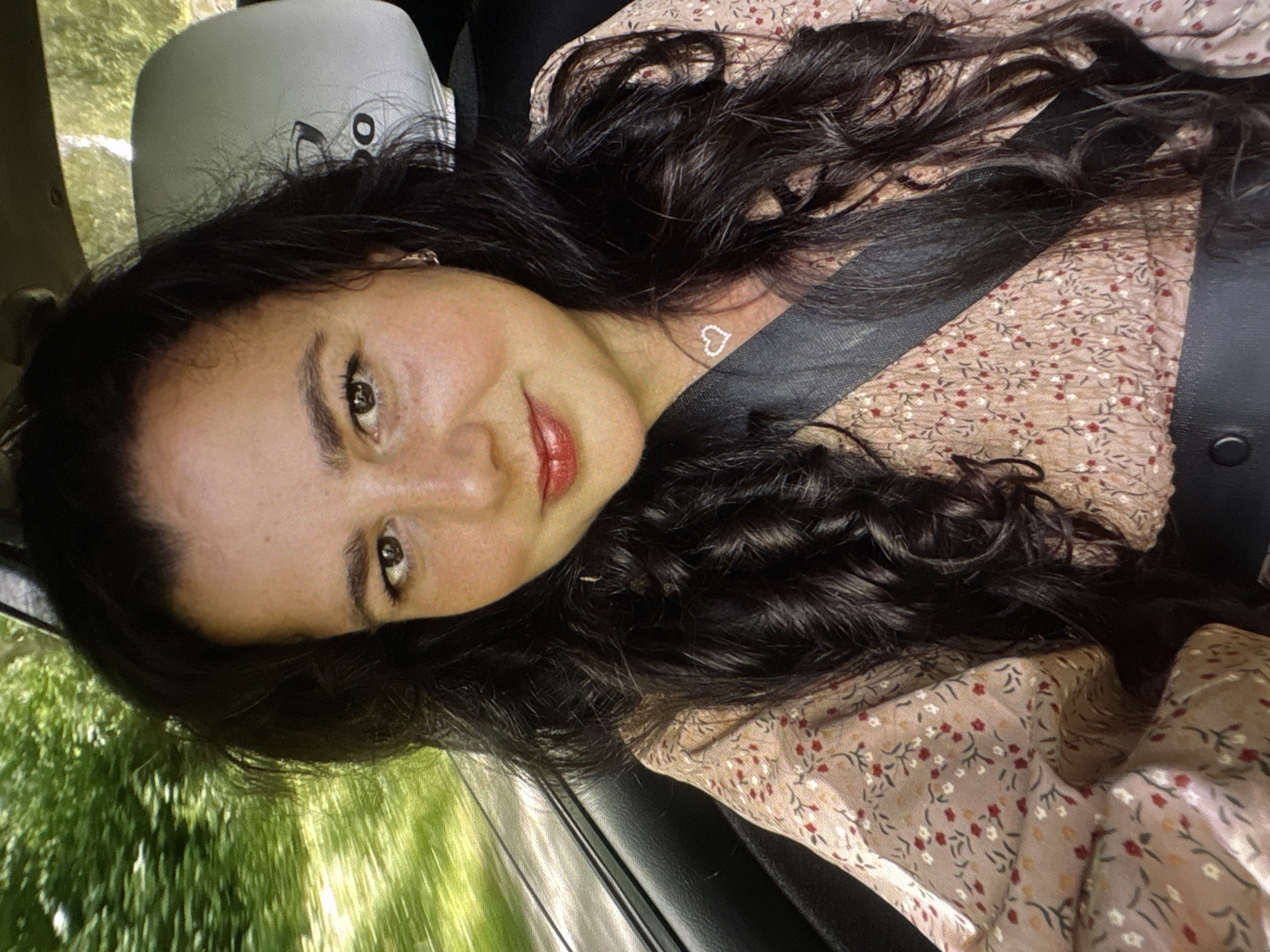 A young woman with long, dark, wavy hair is lying down, possibly in a car, looking at the camera. She is wearing a light-colored, floral print dress and a black seatbelt is visible across her chest. Her hair is spread around her head and shoulders. She has makeup on, including red lipstick, and she is wearing a small, heart-shaped necklace. The background shows a blurred green, leafy scene, suggesting she is in a vehicle moving through a natural environment. The image is taken from a close-up, low-angle perspective.