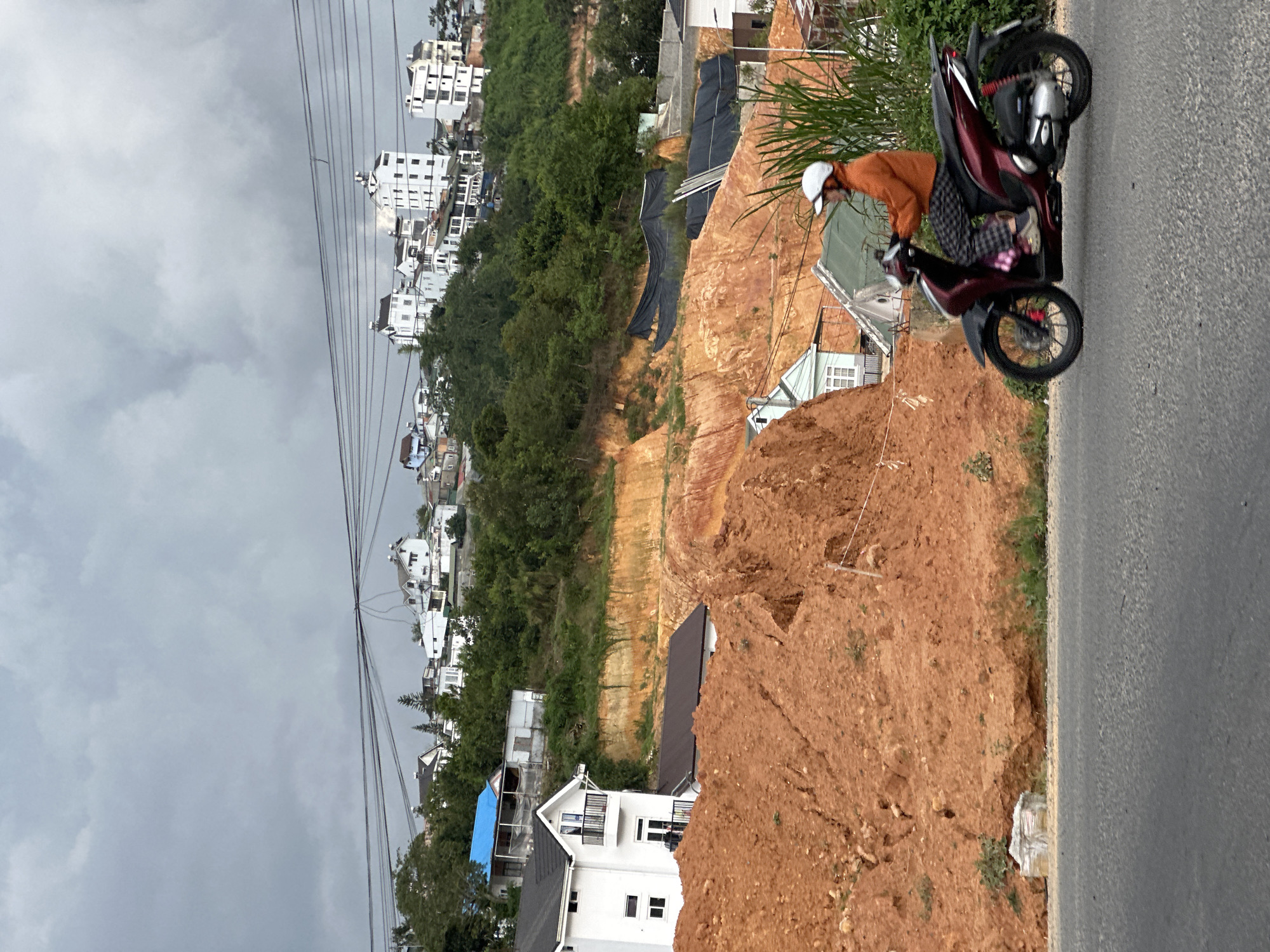 The image captures a scene in Da Lat, Vietnam, featuring a person riding a maroon scooter on a paved road. The rider, wearing an orange jacket and a white helmet, is seated on the scooter with their legs spread. The scooter has black and silver accents and appears to be in motion.
In the background, a significant landslide has occurred, revealing exposed red earth. Several houses are visible on the hillside above the landslide, with a mix of architectural styles, including white buildings with dark roofs and some with blue roofs. Trees and vegetation dot the landscape, with some areas appearing lush and green, while others are barren due to the landslide. Overhead, power lines crisscross the sky, adding to the urban infrastructure visible in the distance. The sky is overcast with gray clouds, suggesting a potentially gloomy or rainy day. The overall scene depicts a rural or semi-urban area undergoing some form of construction or natural event.