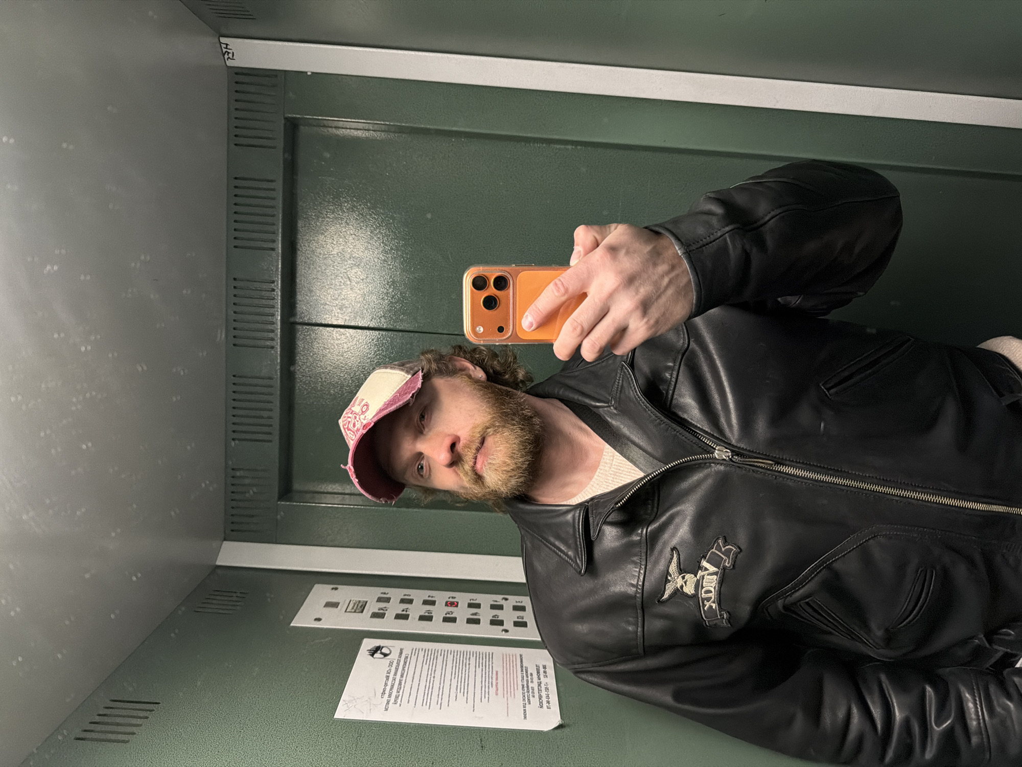 A man is taking a selfie in an elevator. He's wearing a black leather jacket with an embroidered design on the left breast, a pink and white baseball cap, and a light-colored sweater underneath the jacket. He's holding an orange-cased phone. The elevator has a green interior, with button panels and a document posted on the wall. The text on the document, as interpreted by OCR, includes "трио-детям ХО" ООО" and "MOTINI MNORNKADAT NHAПОТАЛ", which suggests the text is in Russian, potentially indicating the city of Saint Petersburg, Russia.