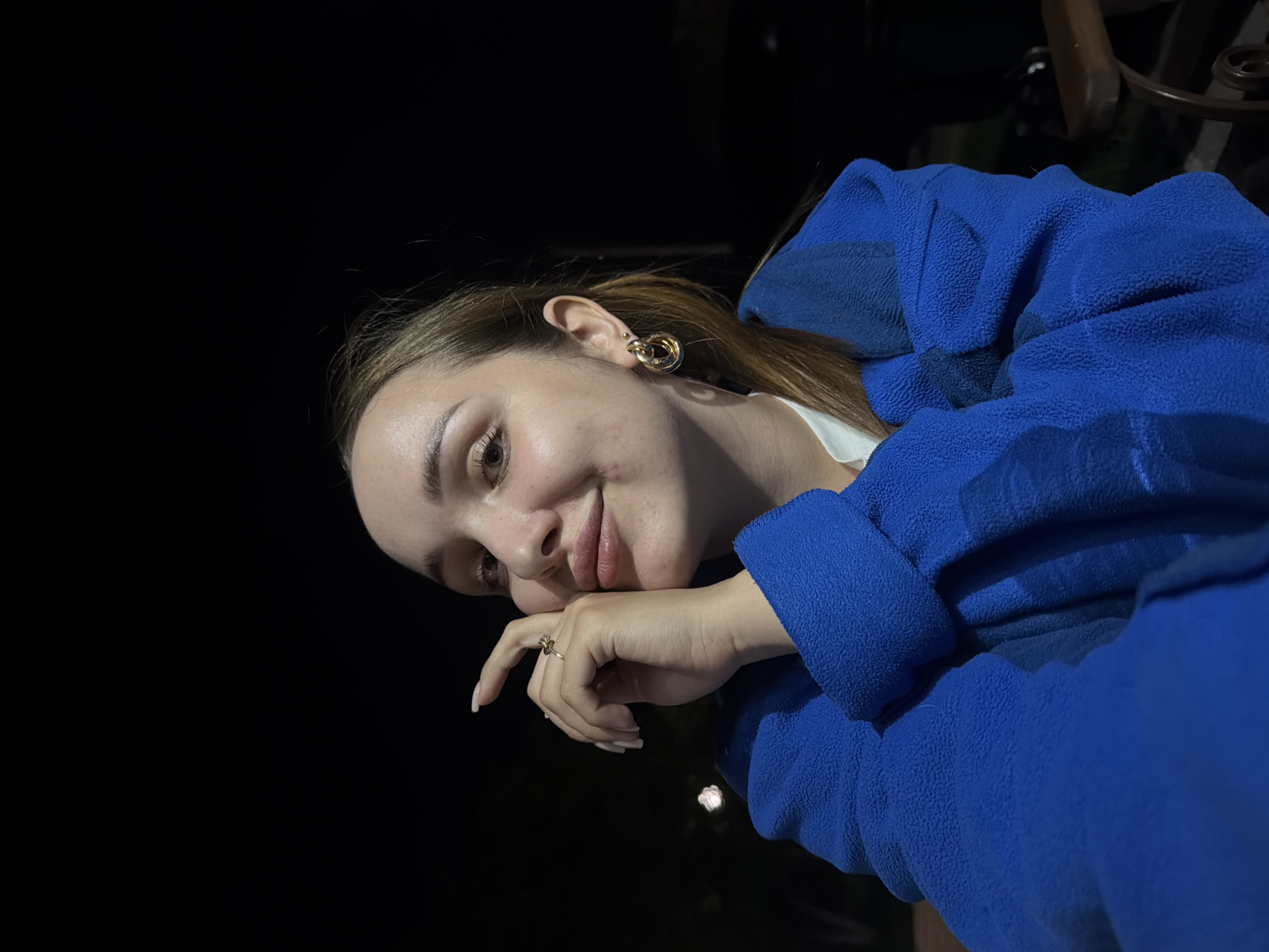 A young woman with fair skin and light brown hair pulled back in a ponytail is posing for a photo. She is wearing gold earrings and a ring. The background is black. The location is unknown.