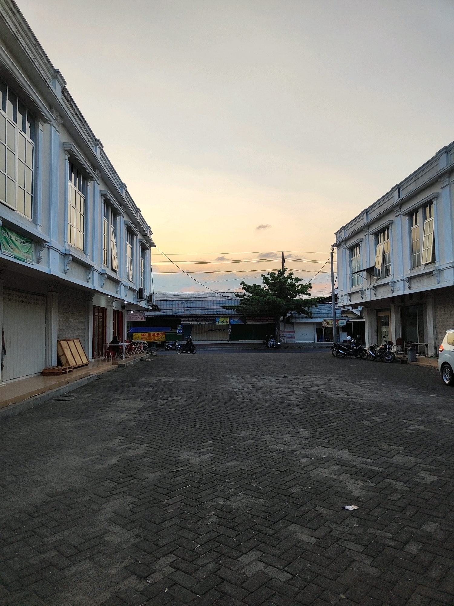 This is a low-angle, full shot of a paved courtyard area in Tamansari, Indonesia, likely in the late afternoon or early evening. Two multi-story buildings with white facades and arched window frames flank the courtyard, creating a symmetrical effect. The building on the left has several closed storefronts on the ground level and signs of commercial activity, including stacked wooden planks. The building on the right also features storefronts.
Several motorcycles are parked in front of the buildings, with two individuals on one of them. A person is seated at a table outside one of the businesses on the left. In the background, beyond the courtyard, there are more structures, a lone tree, and power lines against a pale sky with subtle hues of orange and yellow, indicating sunset. The ground is paved with dark interlocking bricks. A white car is partially visible on the far right. The overall impression is one of a quiet commercial district.
