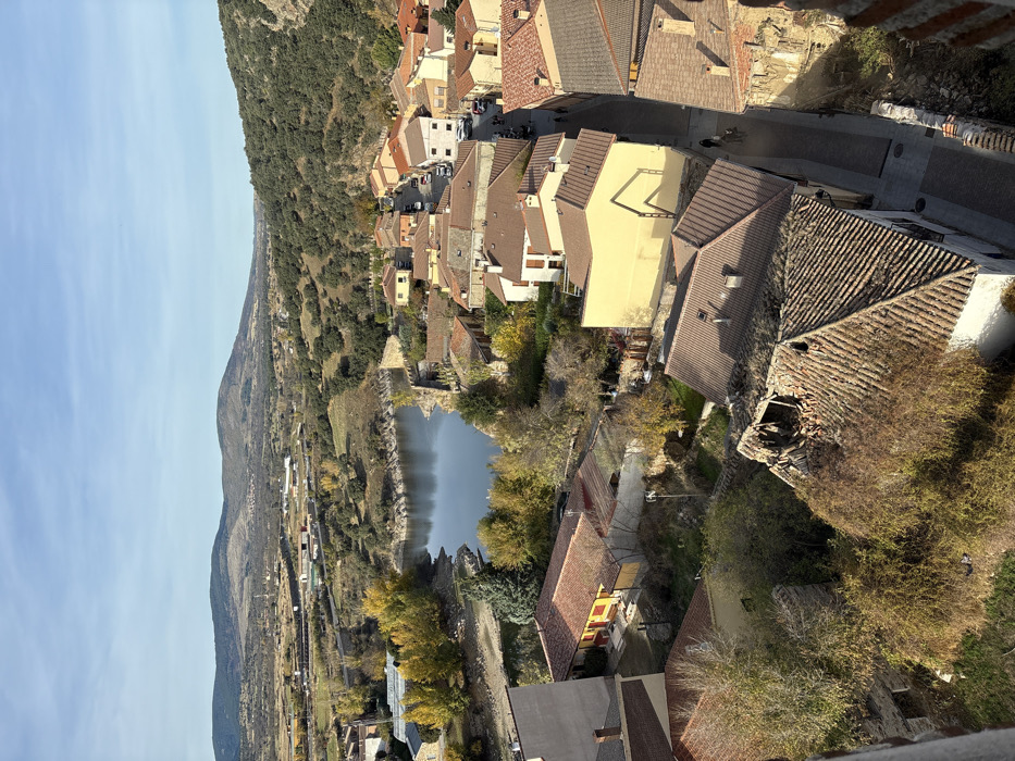This elevated, forensic-style image captures a portion of the town of Buitrago del Lozoya, Spain, under clear, sunny conditions. The scene primarily depicts residential architecture, a body of water, and surrounding natural landscapes.
In the foreground and mid-ground, a dense cluster of buildings with predominantly reddish-brown terracotta tiled roofs is visible. Building facades exhibit varying colors, including beige, yellow, and white. A prominent detail in the lower-mid foreground is a roof with significant structural damage, featuring a large, irregular collapse exposing internal timber framing and debris. Adjacent to this, another roof is extensively overgrown with dense, dried brown vegetation. Narrow, dark-paved streets wind between these structures.
Several parked vehicles are visible on streets in the mid-ground. At least two individuals are discernible walking along a narrow street in the lower-right portion of the image, moving away from the viewer.
To the left and center-mid ground, a calm body of water, likely the Lozoya River or an associated reservoir, is present, reflecting the blue sky. Its banks reveal areas of exposed earth, and stone walls, possibly historical fortifications, line sections of the waterway. Trees with bright yellow autumnal foliage are interspersed throughout the town and along the water's edge.
The background consists of rolling hills and mountains, largely covered with varying densities of green and brown vegetation, extending under a clear blue sky with minimal cloud cover. Distant structures and sparse development are visible in the expansive landscape beyond the town. The viewpoint appears to be from an elevated structure, with a partial brick wall visible in the extreme lower right.