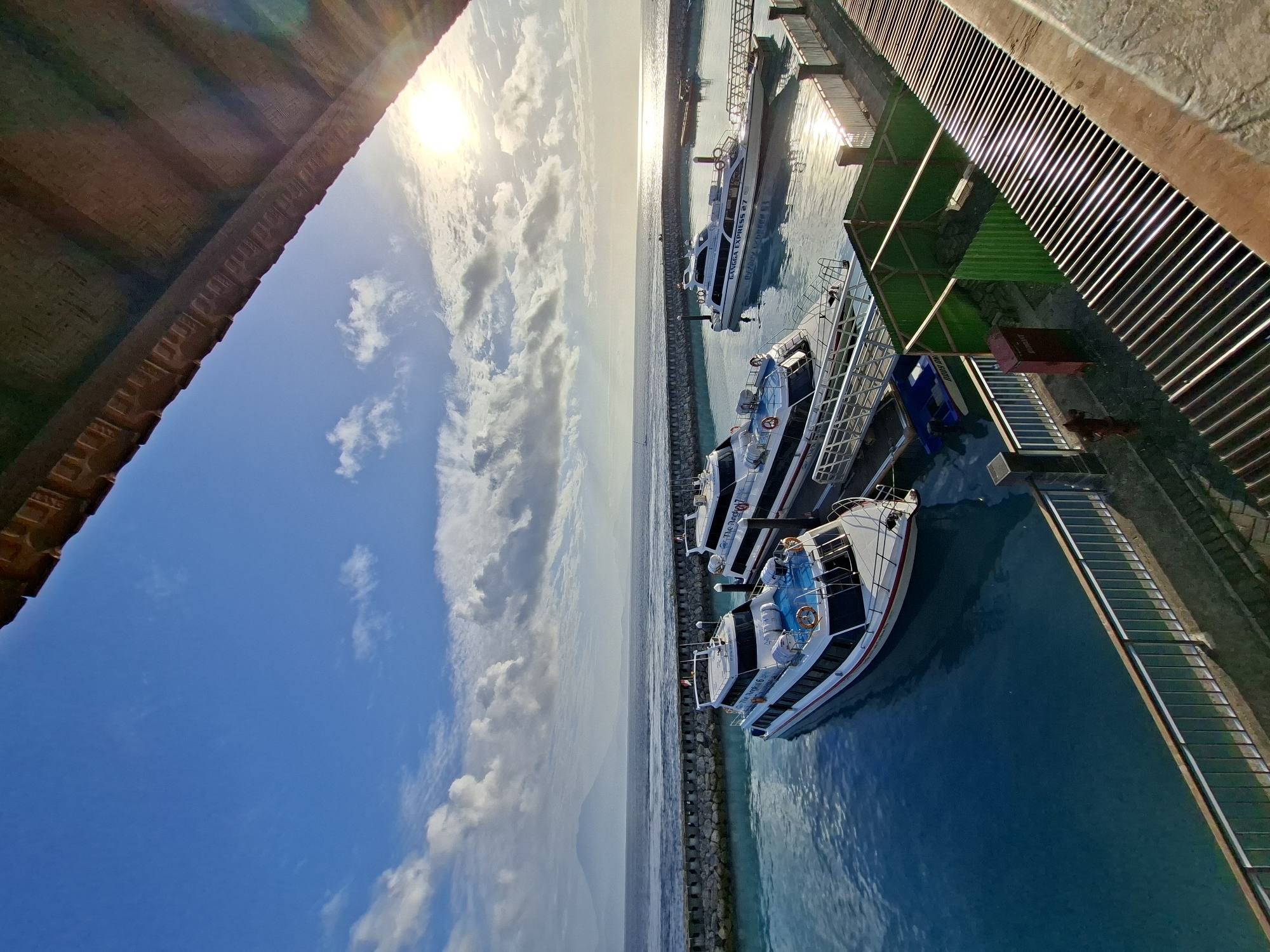 The image shows a harbor on Nusa Penida, Indonesia, with several fast boats docked. The water is clear and blue, and the sky is partly cloudy with the sun shining brightly. The boats are white with red and blue accents. One boat is labeled "GANGGA EXPRESS 27". In the background, a mountain range can be seen in the distance. The foreground shows a walkway with a metal railing, and a portion of a structure with a corrugated roof. The overall scene is serene and picturesque, typical of a tropical island destination.