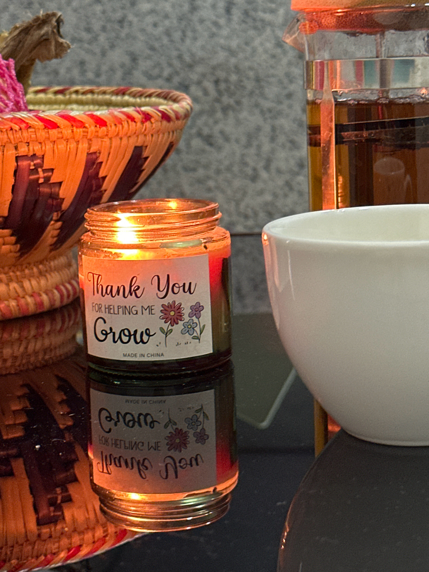 A lit candle in a glass jar sits on a reflective surface. The candle's label reads "Thank You For Helping Me Grow" with floral illustrations. To the left of the candle is a woven basket with red and orange accents, and to the right is a white ceramic cup and a glass carafe containing a dark liquid. The background is a textured gray wall. The scene appears to be an indoor setting, potentially a home or cafe, in Lalitpur, Nepal, characterized by domestic items and a serene ambiance.
