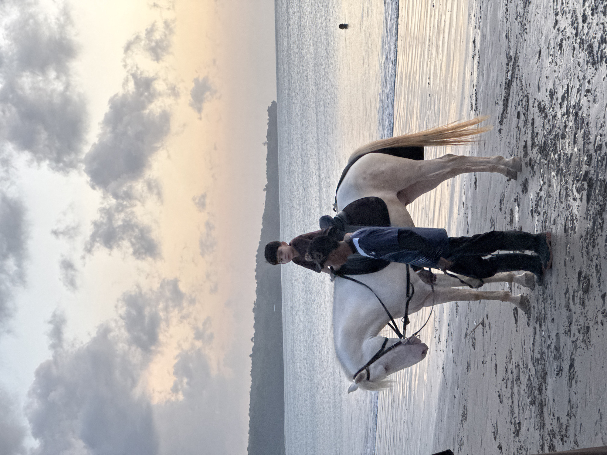 A slightly tilted, medium shot shows a white horse and two people on a beach at sunset in Kuta Selatan, Indonesia. The horse, predominantly white, is saddled and bridled, and stands on the wet, sandy shore with its tail partially submerged in the shallow water. The rider, on the horse's back, is partially obscured by a second person standing beside the horse. This person wears a dark blue shirt and black pants. The background reveals a body of water, likely the ocean, leading to a silhouetted landmass. The sky is filled with clouds, with hues of grey and orange from the setting sun. The overall lighting is soft, suggesting a tranquil, end-of-day scene.