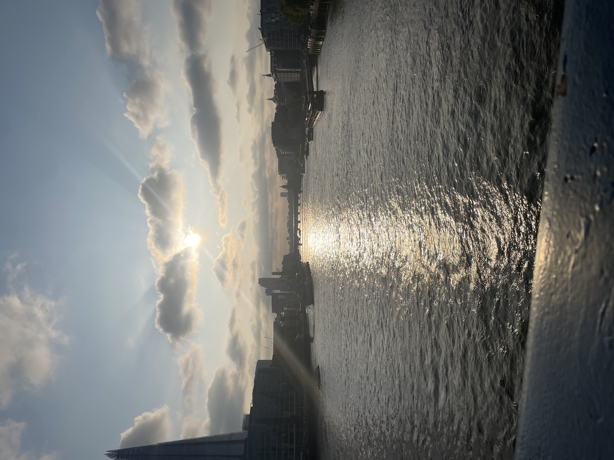The image captures a section of the River Thames in London, United Kingdom, illuminated by a bright sun. The sun, partially obscured by clouds, casts distinct rays into the sky. The river reflects the sunlight, creating a shimmering effect on the water's surface. The cityscape along the riverbank is visible, including buildings, bridges, and cranes. The Shard skyscraper is identifiable on the left side of the frame. The image is taken from an elevated position, possibly from a bridge. A portion of the structure from which the photo was taken is visible in the lower right corner.