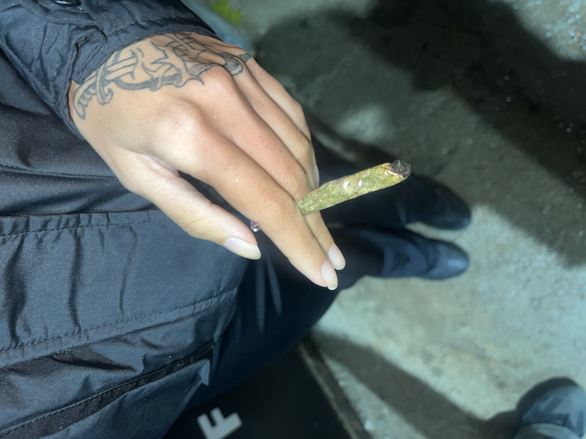 A person is holding a lit marijuana cigarette in their left hand. The person is wearing a black jacket and has a tattoo on the back of their hand. They appear to be sitting. The background is an out-of-focus concrete surface. It is not possible to determine if this photo was taken in Lomas de Zamora, Argentina or any specific location.