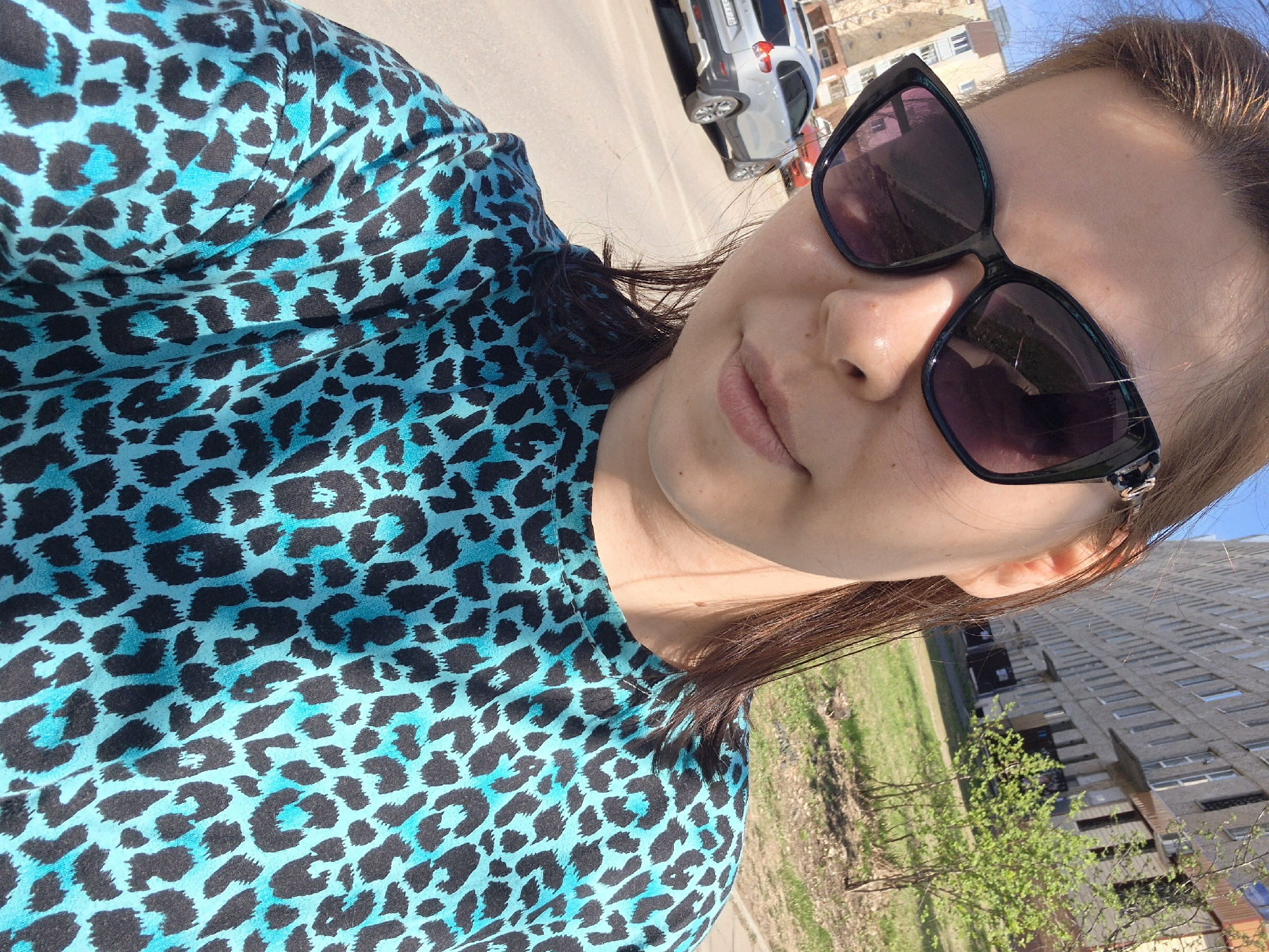 A woman with light skin and brown hair, wearing black sunglasses and a blue and black leopard-print shirt, is taking a selfie outdoors. In the background, there is a light-colored building with multiple floors and windows, suggesting an apartment complex. Patches of green grass and small trees are visible in front of the building. A road runs alongside the building, with a silver car parked on it. The image appears to be taken in the city of Tobolsk, Russia.