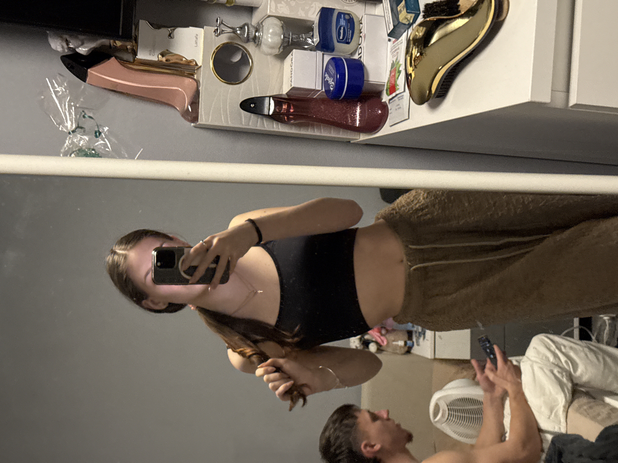The image appears to be a mirrored selfie taken in an indoor environment, likely a bedroom or bathroom. A woman is holding a smartphone, positioned to capture her reflection in a mirror. She is wearing a black crop top and brown pants. In the reflection, a man is lying on a bed, operating a remote control. A white fan is visible on the floor near the bed. The background behind the mirror shows a cluttered shelf with various items including toiletries, hair accessories and boxes. The location in Sofia, Bulgaria cannot be determined from the given image.