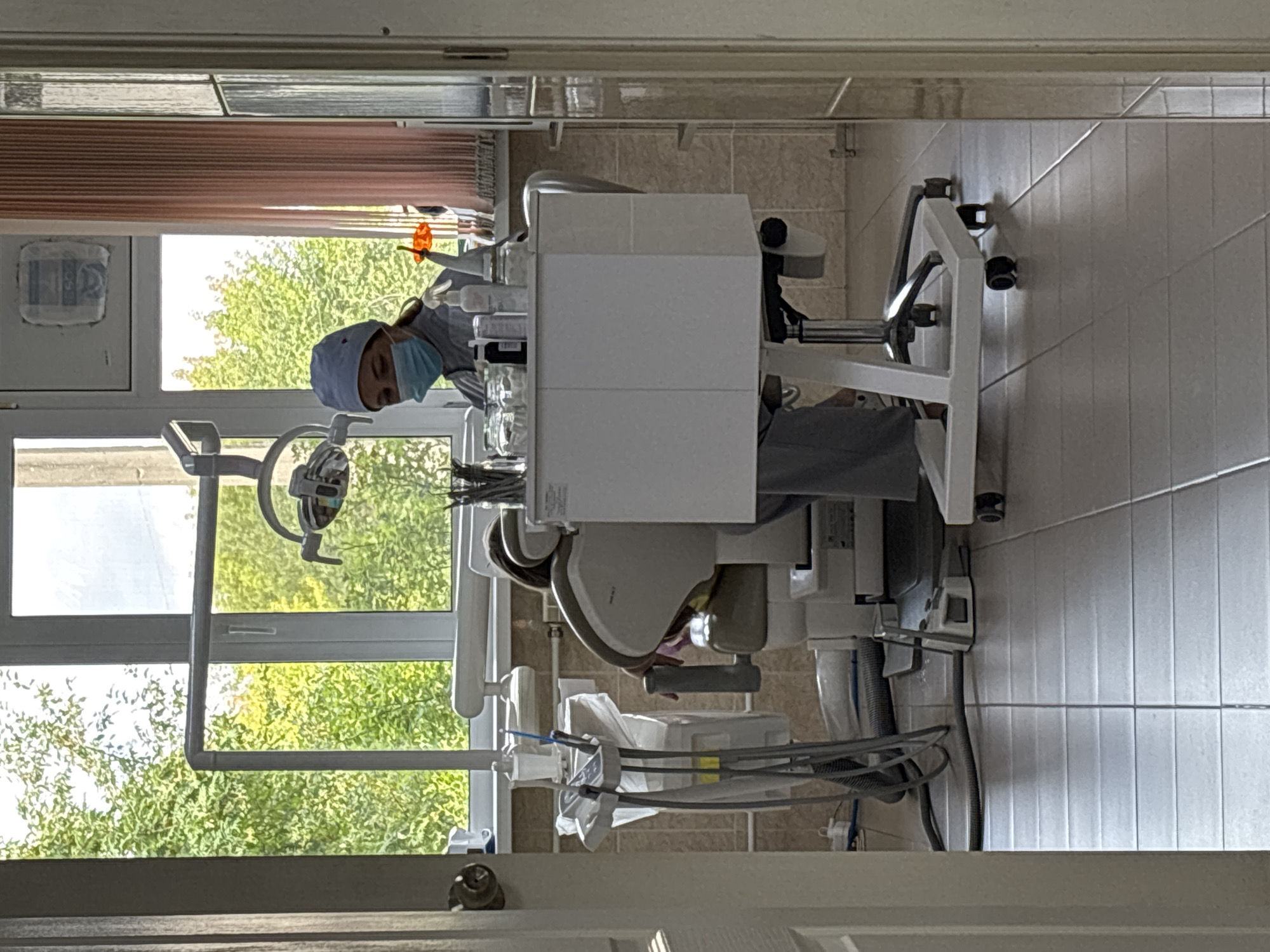 The image depicts the interior of a dental operatory, likely in Artemovsky, Russia. A female dentist, wearing a light blue surgical cap and mask, is seated behind a white dental cart. She appears to be engaged in a procedure, leaning forward towards a patient who is seated in a beige and gray dental chair. The patient's head is not fully visible.
In the background, a large window looks out onto lush green trees, suggesting the operatory is located on a lower floor or has an unobstructed view of outdoor foliage. A dental examination light is suspended above the patient chair. Various dental instruments and supplies are visible on the cart, including jars, bottles, and what appear to be surgical tools. The floor is tiled with light-colored square tiles. The room has beige tiled walls. A pink pleated curtain is visible on the right side of the window. The overall impression is a clean, functional medical environment.