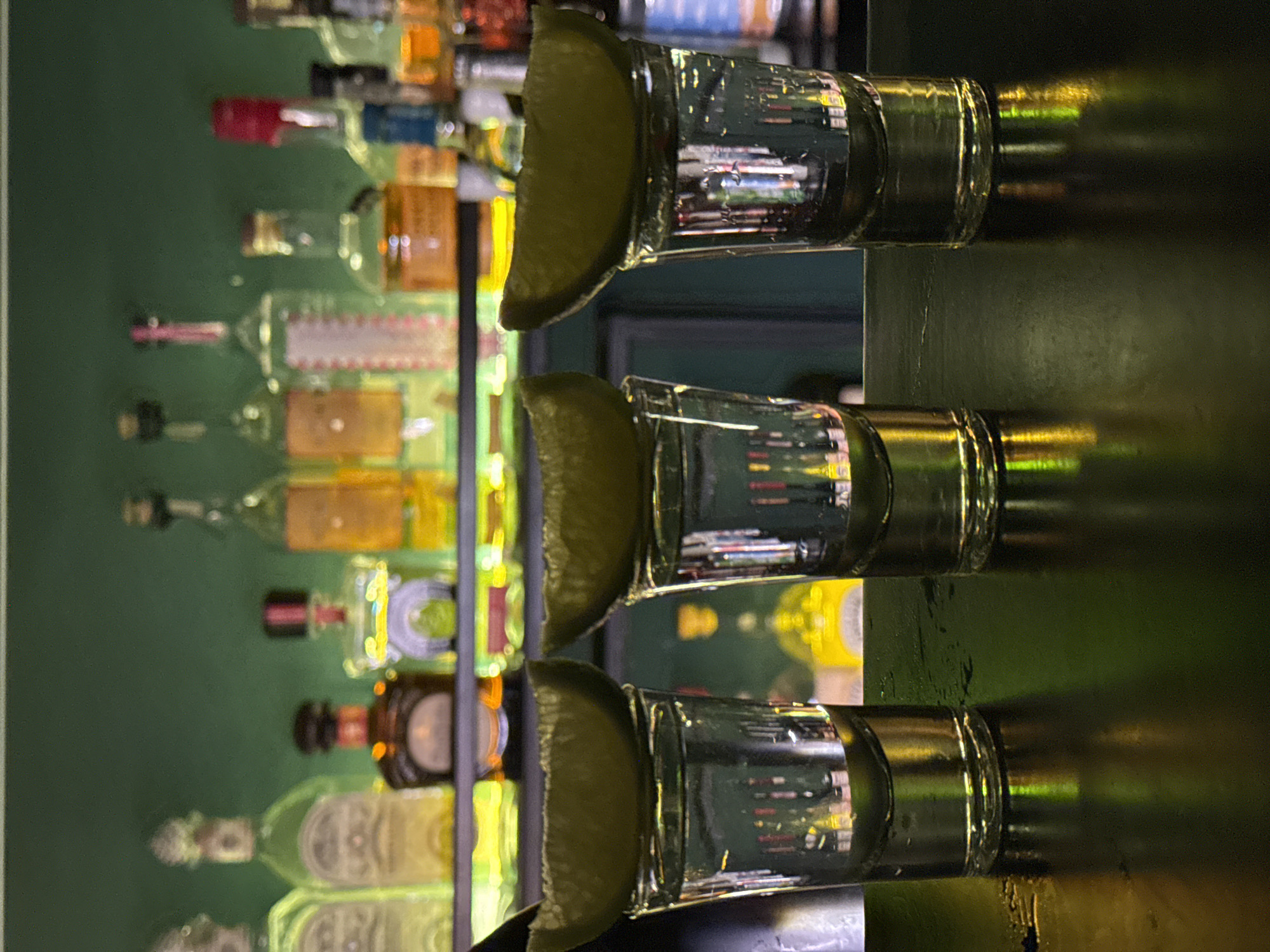 Three shot glasses with lime wedges are aligned horizontally on a surface with a reflective sheen. Behind the shots, blurred bottles are visible in the background, likely on a bar shelf. The bottles are various shapes and colors, suggesting a variety of alcoholic beverages. The background is green. The lighting is dim. This is likely a scene in a bar. Budva, Montenegro is not discernible from the image.