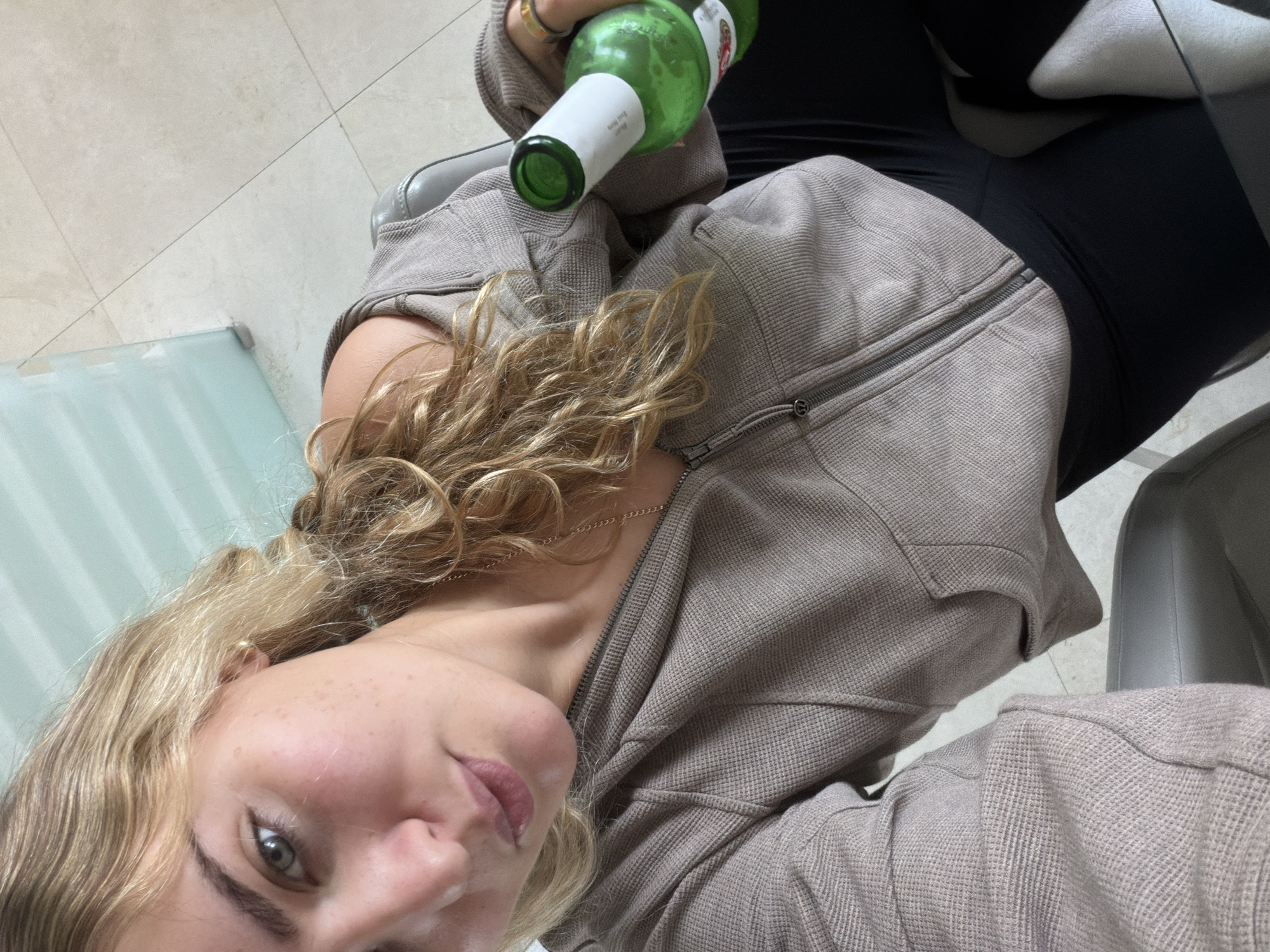 A young woman with blonde, curly hair and freckles is seated and angled towards the camera, holding a green glass bottle. She is wearing a brown zip-up hoodie over black leggings. The setting is indoors, possibly a room in a residence or establishment in Key Biscayne, United States of America, evident by the tile flooring and hint of a glass table or wall. The floor is beige tile and there is a glimpse of a chair.