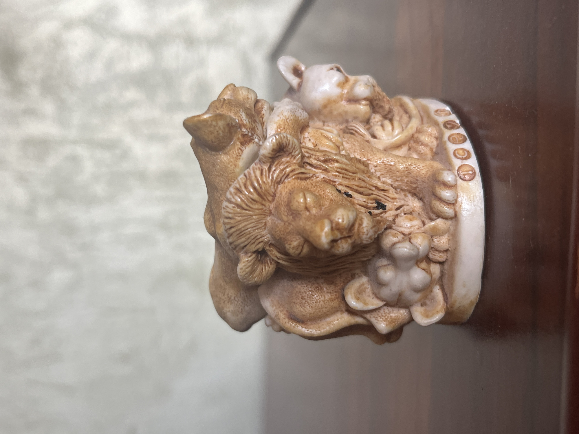 A small, tan-colored figurine depicts a lion with a thick mane, surrounded by two smaller lions or lionesses. The main lion is in the center, with its eyes closed and head tilted slightly forward. One smaller lion is lying on its back, its head resting on the main lion's mane, while another is positioned to the right, appearing to look away. The figurine is placed on a dark brown wooden surface. The background is a light-colored, textured wall. There are no people present, and no specific location in Vladimirovka, Russia is identifiable.