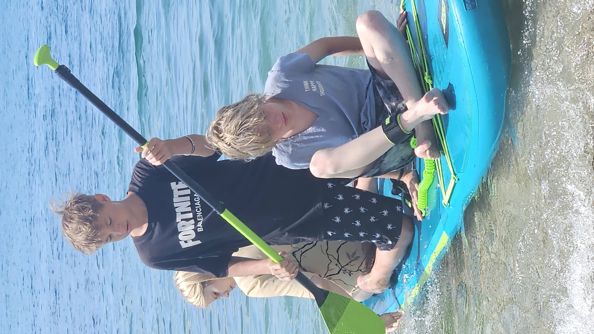 The image captures a bright, sunny day at the beach in Novomikhaylovsky, Russia. Three young individuals are positioned on a turquoise paddleboard in the clear, shallow, blue water. The individual in the foreground, a blond boy wearing a grey t-shirt with the text "THINK HAPPY THOUGHTS" and patterned shorts, is seated with his legs crossed. He has a black ankle strap around his right ankle. To his left, another boy, also with blond hair, stands holding a paddle. He is wearing a black t-shirt with "FORTNITE BALENCIAGA" printed on it and black shorts with white palm tree patterns. Behind them, a young person with blond hair, partially visible, is seated, wearing a light-colored shirt. The paddleboard is equipped with green cords. The water is calm, with small ripples around the paddleboard, and the seabed is visible through the clear water. The sky above is a vibrant blue.