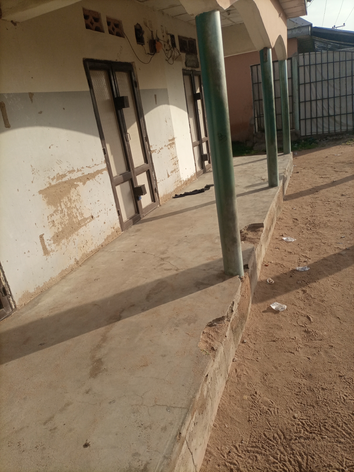 The image shows the exterior of a building with a covered walkway supported by several green, cylindrical pillars. The building's facade is weathered, with peeling paint and visible brickwork in some areas. There are several doors along the walkway, some with glass panels. The walkway itself is concrete and appears to be in a state of disrepair, with cracks and crumbling edges. The ground beside the walkway is sandy and uneven, with some litter scattered on it. In the background, a metal fence is visible. The scene appears to be in Jalingo, Nigeria.