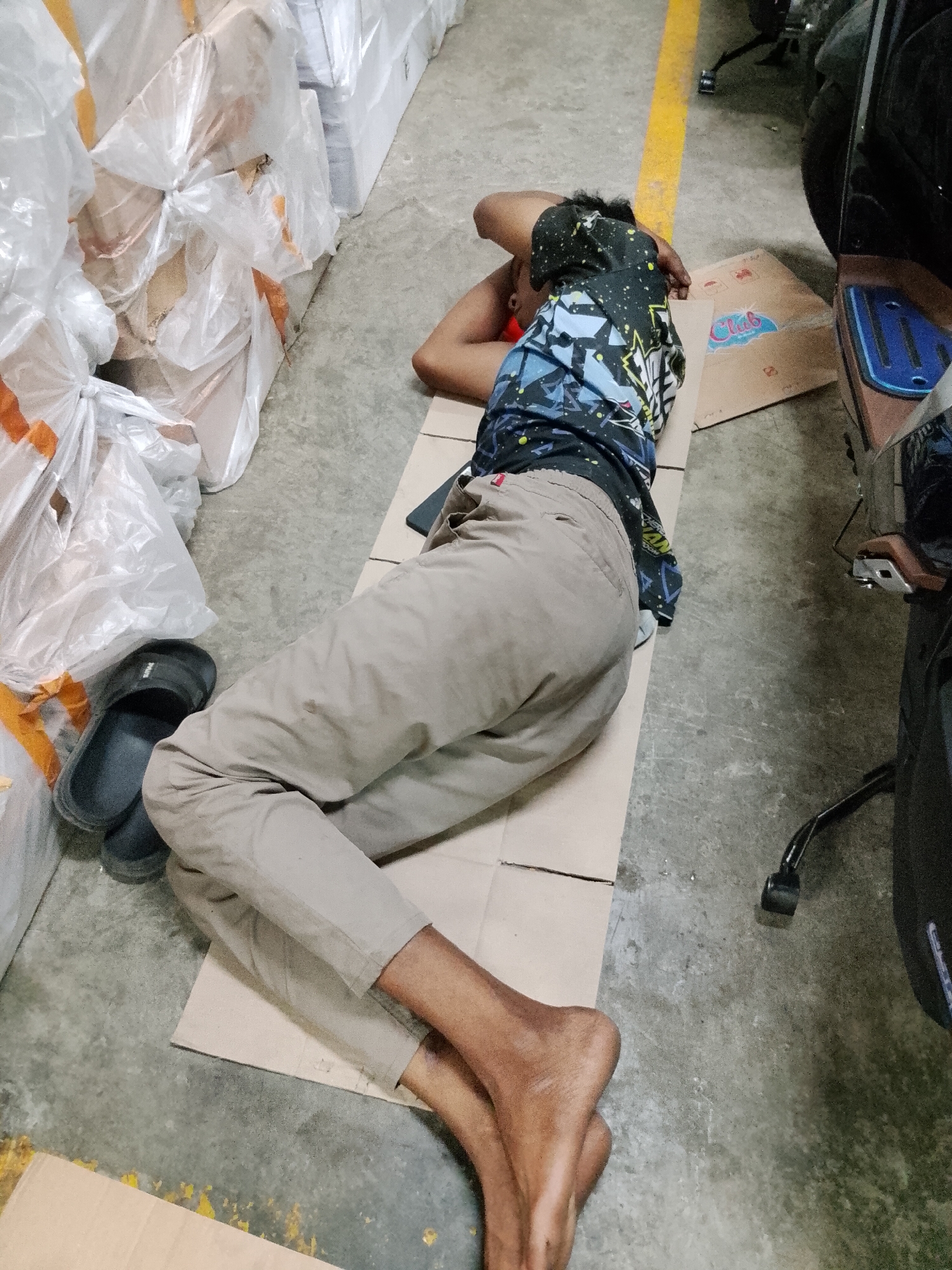 An adult male is lying asleep on a cardboard on the floor of a warehouse, possibly in the city of Pasuruan, Indonesia. He is wearing a black t-shirt with a colorful graphic design and light brown pants. His left arm is tucked under his head, and his right arm is resting on his chest. His legs are crossed at the ankles. To his left are several large plastic-wrapped packages, and to his right is a dark-colored motorcycle. A yellow line is painted on the floor behind him. The floor is concrete and appears to be dirty. A pair of black sandals is placed near the packages.