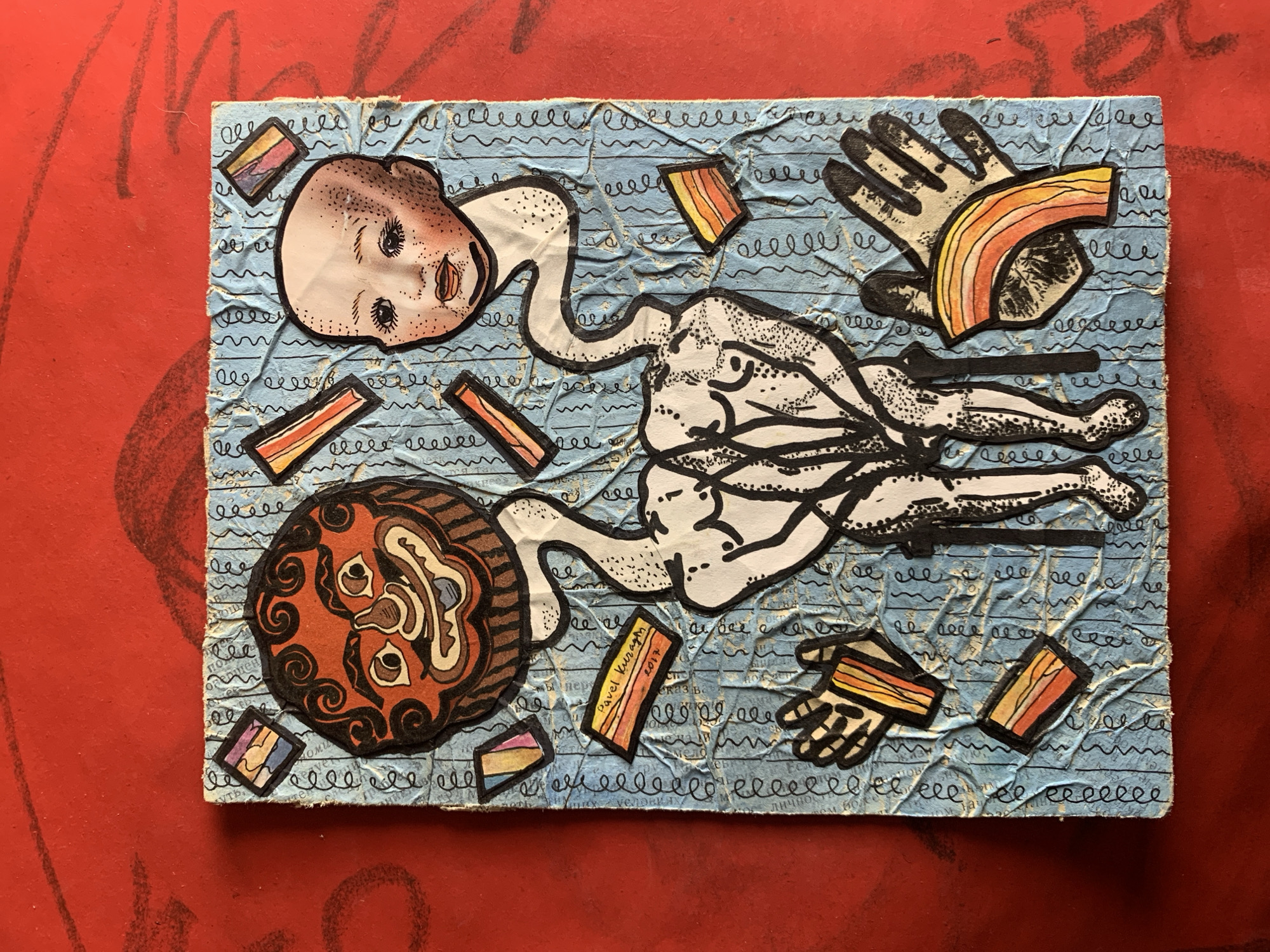 The image shows a piece of mixed-media art on a bright red, textured surface. The artwork itself is a collage on a light blue, crumpled background covered with black squiggly lines.
Dominating the center of the artwork is a stylized, anatomical torso rendered in black and white ink. The torso appears to be composed of two figures intertwined or fused together. Attached to the top of the torso are two distinct heads. On the left is a mask-like face with an exaggerated, grinning expression, featuring large eyes, a wide mouth, and curly black hair. On the right is a pale, disembodied baby's head with wide-set eyes, rosy cheeks, and freckles.
Scattered around the central figures are various small, colorful rectangular and diamond shapes with rainbow-like or striped patterns. To the left of the torso, there are two black and white drawings of hands. One hand is open with fingers spread, and the other is slightly curled. To the right of the torso, there is another drawing of an open hand, with a colorful, arched shape overlaying it.
A small orange rectangle in the lower left quadrant of the artwork contains handwritten text. It reads "Pavel Kuragin" and "2017".
The artwork is positioned on a flat surface with a prominent red texture, possibly a table or floor, with visible brush strokes or patterns. The lighting appears to be overhead, casting soft shadows.
The image is a photograph of the artwork and does not depict any specific location within Sergeevo, Russia, nor does it contain any information or context related to that city.