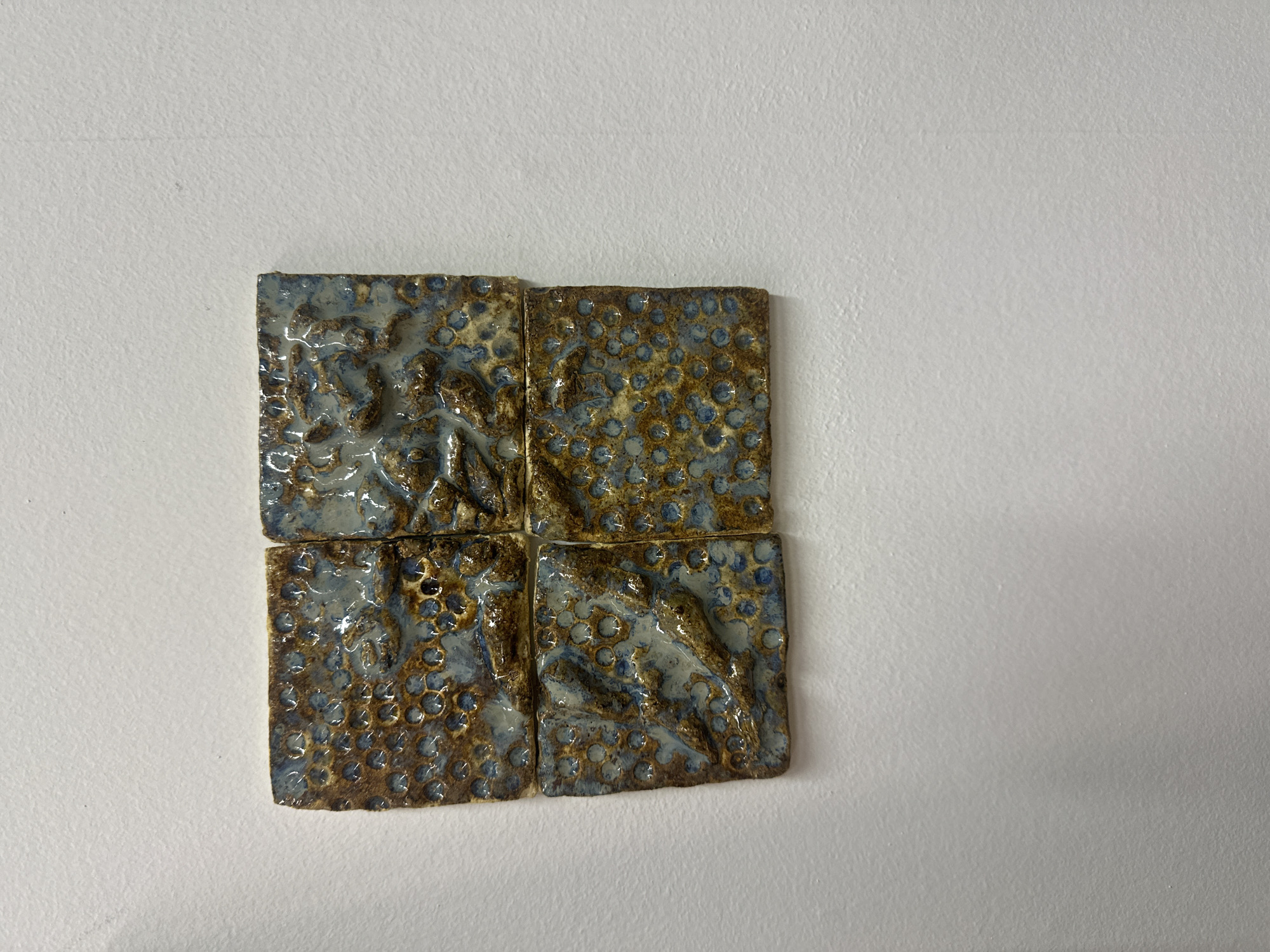 Four square, textured tiles are arranged in a 2x2 grid on a plain white surface. The tiles have a glaze with a mix of brown and blue-grey colors. The glaze is uneven and textured, with some areas appearing bubbled or cratered. The surface appears to be a piece of paper or a similar material. There is no indication of a location in Moscow, Russia or any people in the scene.
