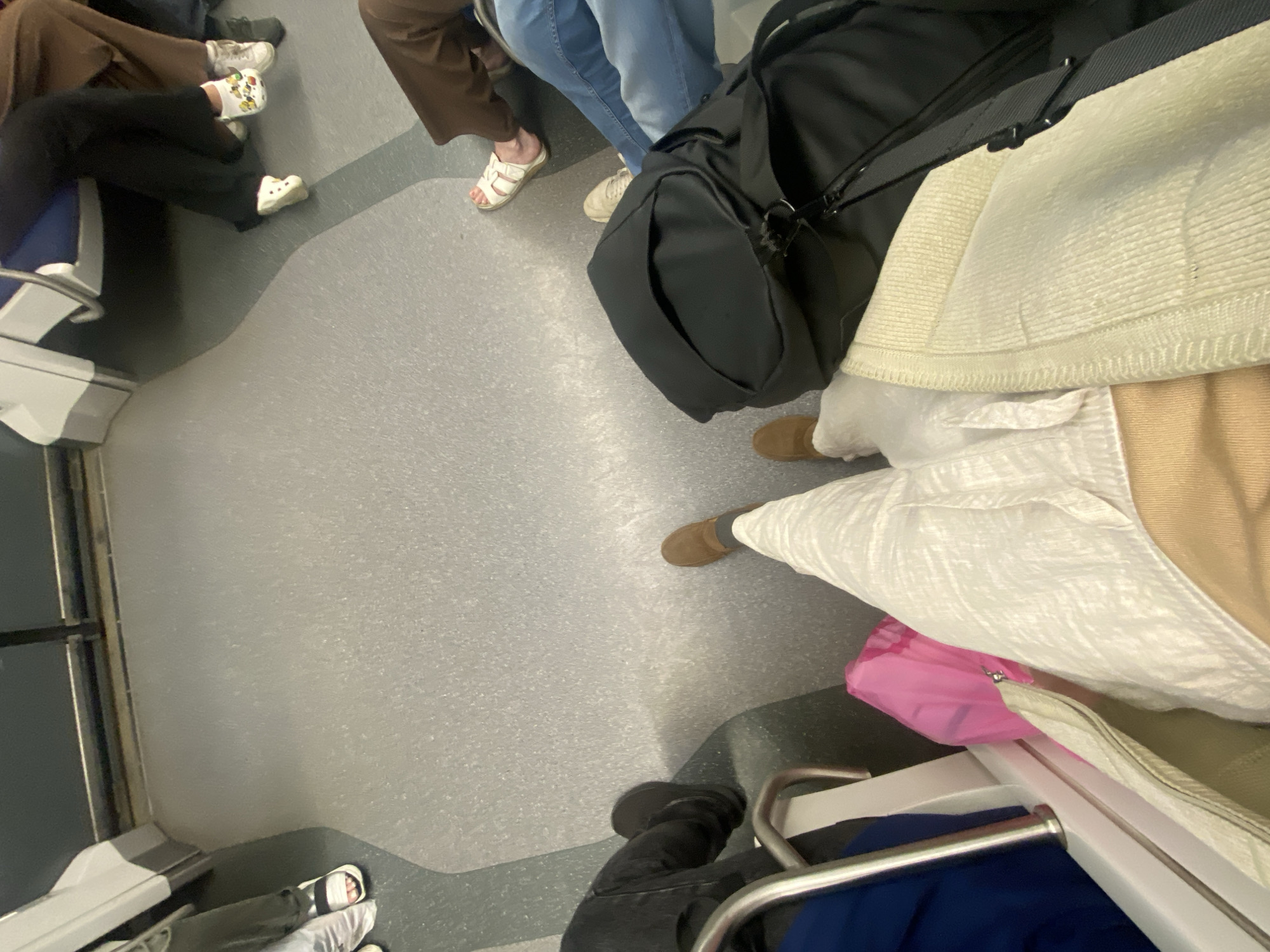 The image depicts the interior of a subway car. Several individuals are seated or standing, their legs and feet visible. One person is wearing white sandals with black straps. Another person wears white Crocs adorned with cartoon characters. A third person is wearing white sandals. A black backpack is visible, placed on the floor next to a person's legs. A pink plastic bag is also present. The floor of the subway car is gray and textured. The lighting suggests an indoor environment, characteristic of public transportation. There is no specific information in the image to indicate the location as Moscow, Russia.