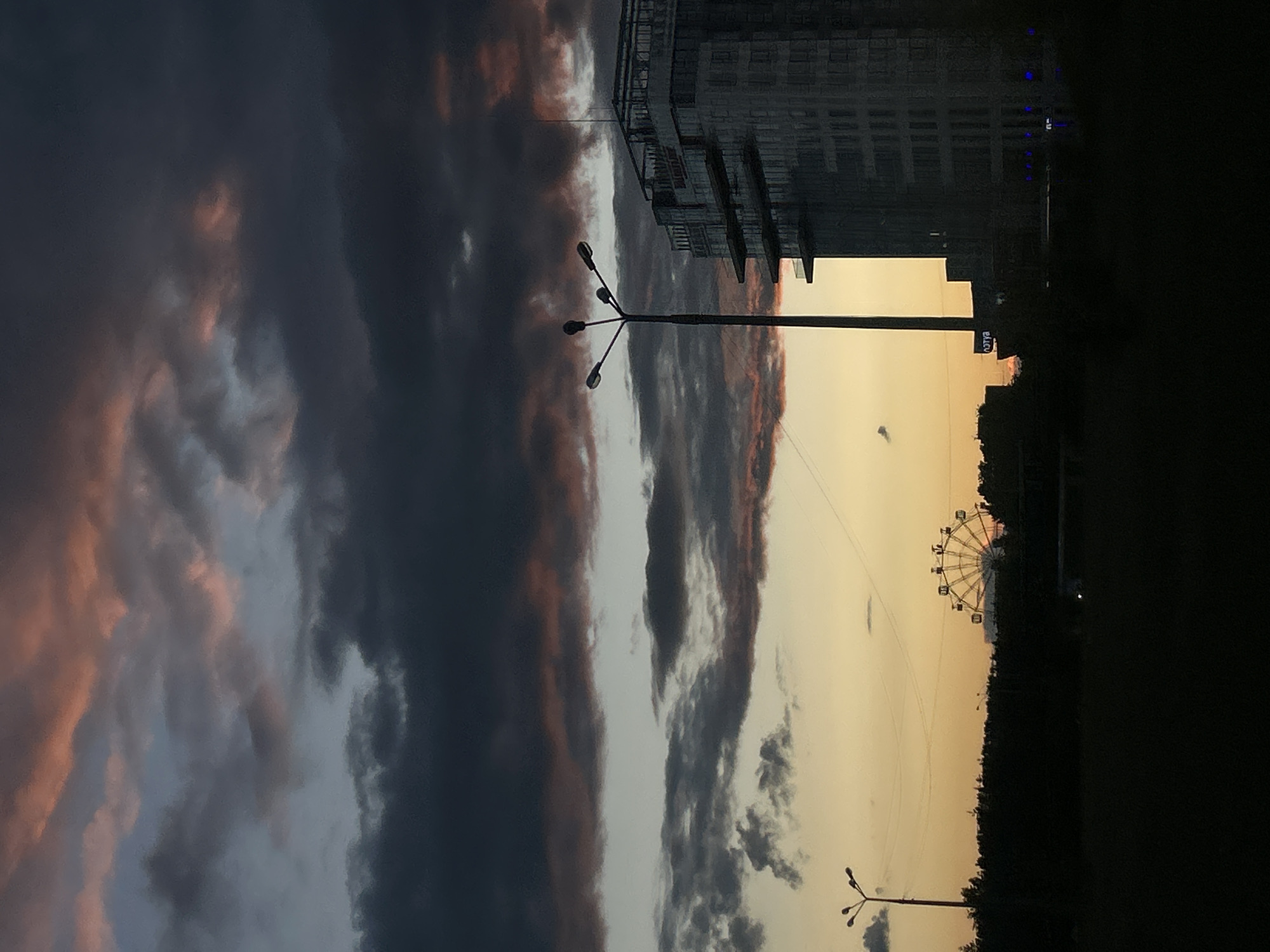The image depicts an urban landscape in Tolyatti, Russia at sunset. The sky is filled with dramatic clouds, with varying shades of grey and pink hues. A tall building is visible on the right side. There is a Ferris wheel in the background and a lamppost in the foreground. A street is visible in the middle.
