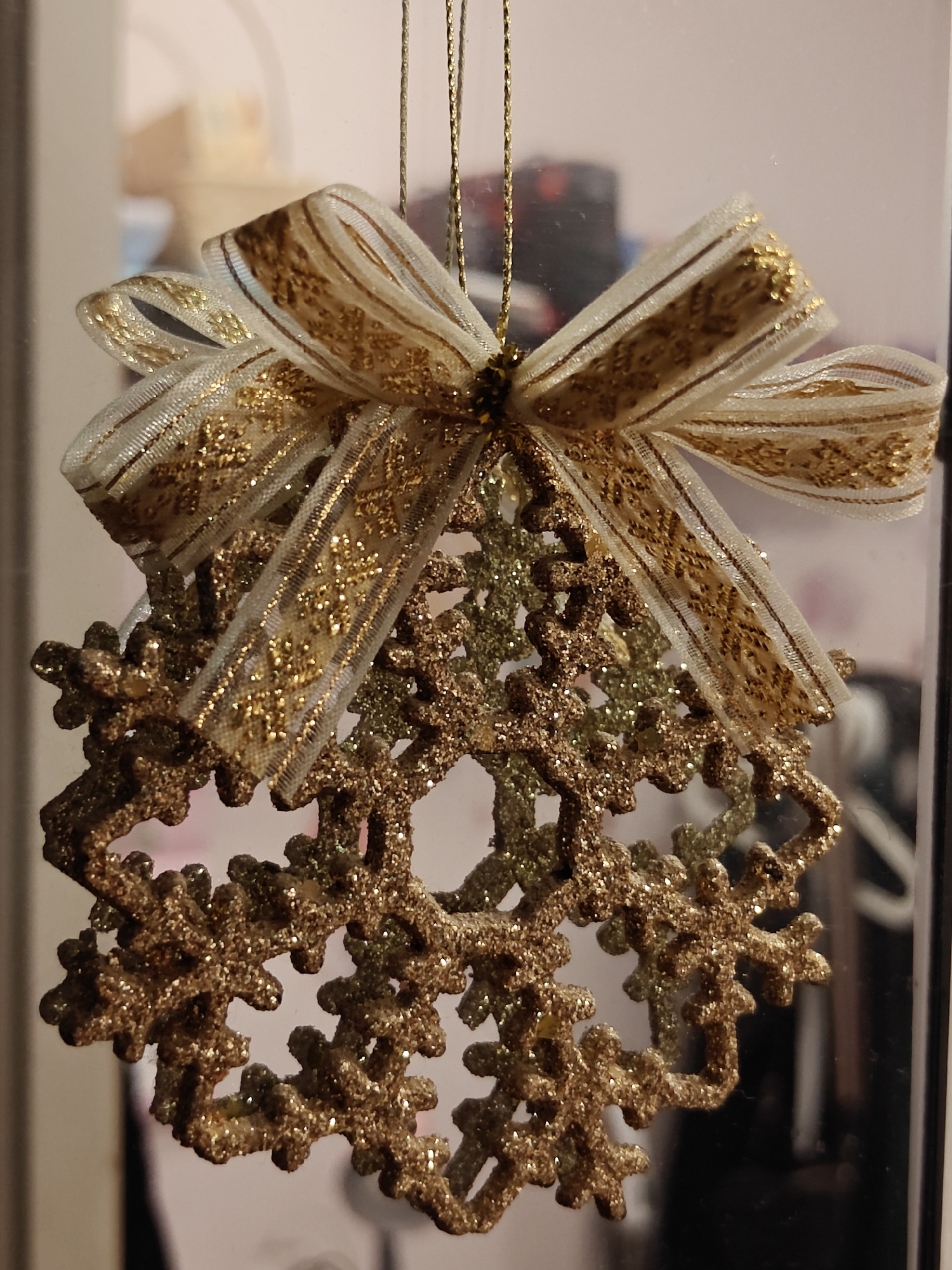 This is a close-up, vertical shot of a Christmas ornament hanging indoors, likely in Saint Petersburg, Russia. The ornament is a golden snowflake, intricately detailed and covered in glitter. It is attached to a thin gold string and topped with a festive bow made of white and gold ribbon, also adorned with glitter. The background is blurred, but appears to be a window or reflective surface, showing indistinct shapes and colors that suggest an indoor environment, possibly a room or a shop window. The lighting is soft, highlighting the sparkle of the ornament. There are no people or discernible actions in the image.