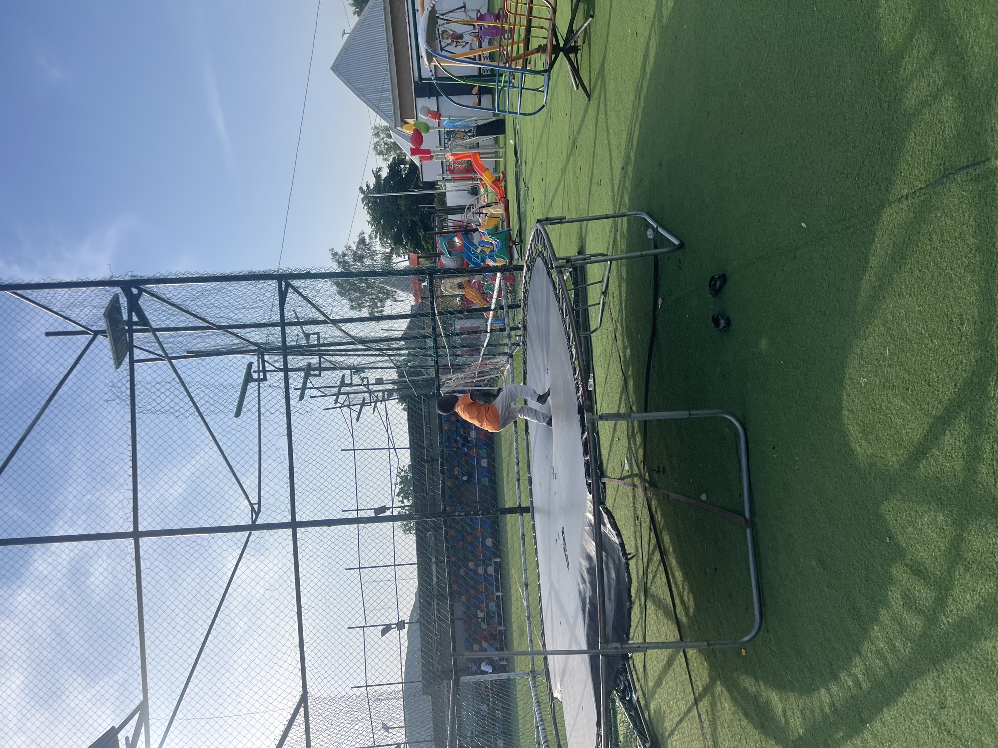 In Jalingo, Nigeria, a young boy in an orange shirt and white pants is captured mid-jump on a large, grey trampoline with a black mat, situated on a bright green artificial turf field. The trampoline is surrounded by a metal frame and netting. In the background, a children's playground is visible, featuring colorful slides, swings, and other play structures. Behind the playground, bleacher-style seating with multi-colored seats is present, suggesting a sporting or recreational facility. The scene is bathed in sunlight, casting shadows on the green field, with a clear blue sky overhead and some light clouds. The overall impression is a vibrant outdoor recreational area.