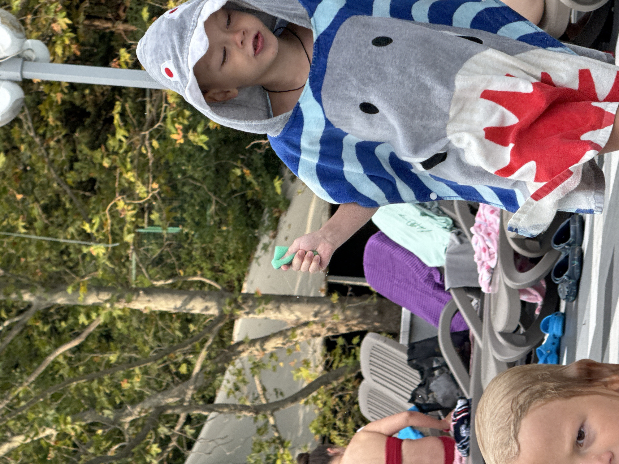 The image shows a young boy wearing a shark-themed hooded towel. He has his eyes closed and his mouth slightly open, as if he is playing or being sprayed with water. He is holding a green sponge in his right hand, from which a thin stream of water is visible. The towel is grey with blue stripes and has shark facial features printed on the hood. In the background, there are trees with green foliage, suggesting an outdoor setting. Partially visible in the background are lounge chairs and some clothing items, indicating a relaxed, possibly poolside or beach-like environment. Another child's head, with blonde hair, is visible in the lower right corner of the image. The overall impression is of a warm day and a child enjoying themselves. The image is taken in Yalta, Ukraine.