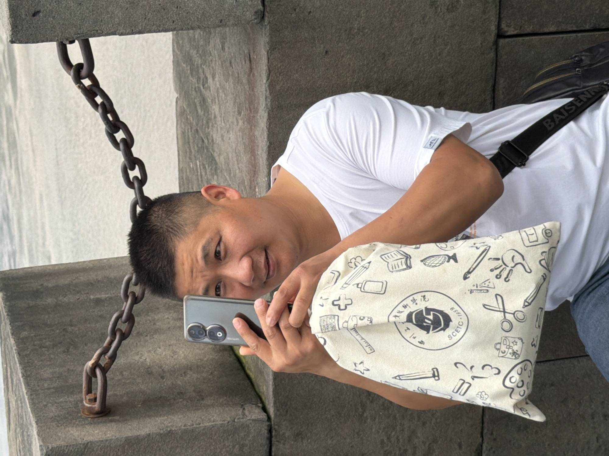 A man of East Asian descent with a buzz cut and a white t-shirt is holding a smartphone up with his right hand, taking a photo. His left hand is holding a canvas bag with a black strap that says "BAISEN" in white letters. The bag is white and covered in a black line-drawing pattern of various objects, including scissors, pencils, and a book. The man is looking at his phone. He is standing in front of a textured concrete wall, which appears to be part of a seawall or embankment, with a thick metal chain bolted to it. The background is out of focus and shows a body of water. The scene appears to be outdoors, possibly along the waterfront in Yuzhong District, China, given the mention of the location.