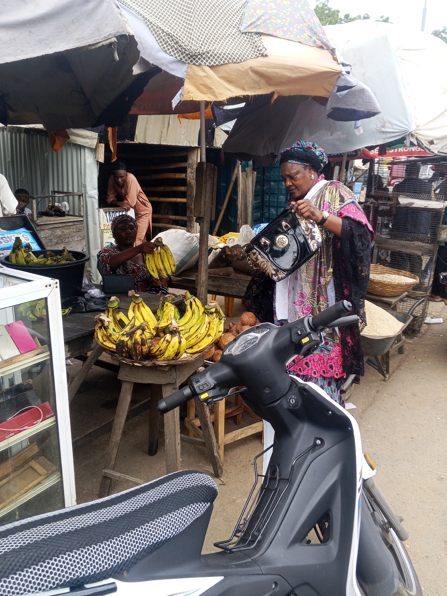 The image depicts a bustling outdoor market scene in Okene, Nigeria. In the foreground, the front of a white and black motorcycle is visible, with its handlebars and front wheel slightly blurred due to the close-up perspective. Behind the motorcycle, a woman wearing a colorful patterned dress and a headwrap is standing, holding a black handbag with gold embellishments. She appears to be interacting with a stall.
To the woman's left, a large black bucket is filled with green bananas, and a round tray overflowing with ripe yellow bananas rests on a wooden table. Another woman, dressed in a tan outfit, is partially visible behind a corrugated metal structure, leaning out and looking towards the market. To the far left, a display case with shelves and some items, possibly electronics, is present. In the background, a man is seen standing near a cage-like structure, and stacks of grain are visible in a wheelbarrow and a basket. The market appears to be set up under tarpaulin shelters, suggesting a permanent or semi-permanent stall area. The ground is covered in dirt and sand. The overall impression is one of a vibrant, lively marketplace common in Nigerian cities.