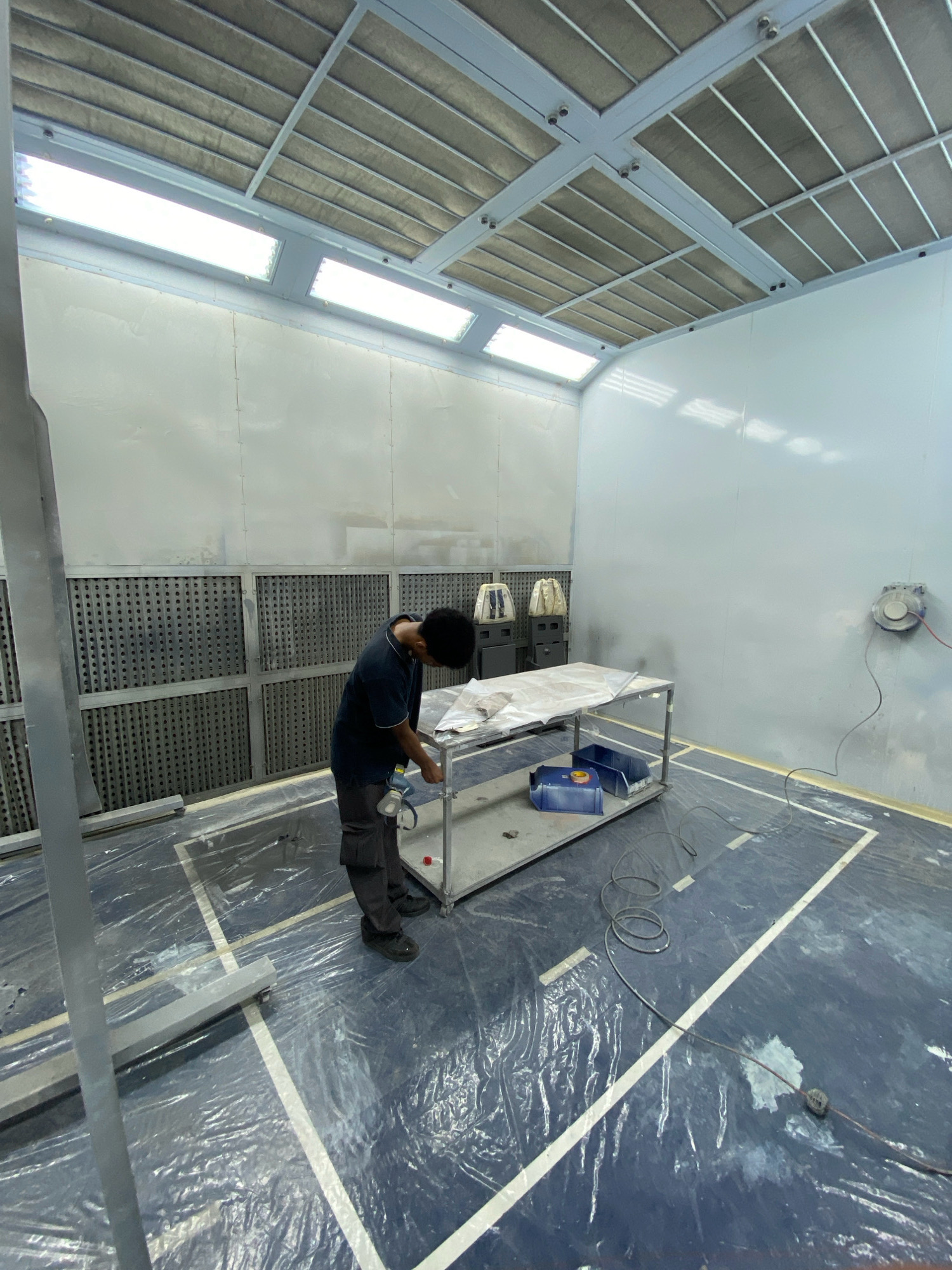 In a spray booth facility in Batam, Indonesia, a young man wearing a dark t-shirt and grey trousers is working on a trolley-mounted table. The room is well-lit with fluorescent lights on the ceiling and appears to be a clean, controlled environment, likely for painting or finishing. The floor is covered in blue protective sheeting, with white lines marking a designated work area. The man is focused on the table, which is draped with a light-colored sheet, suggesting he is preparing an object for painting or has just finished. In the background, industrial equipment, including what appear to be ventilation units and a spray gun, are visible.