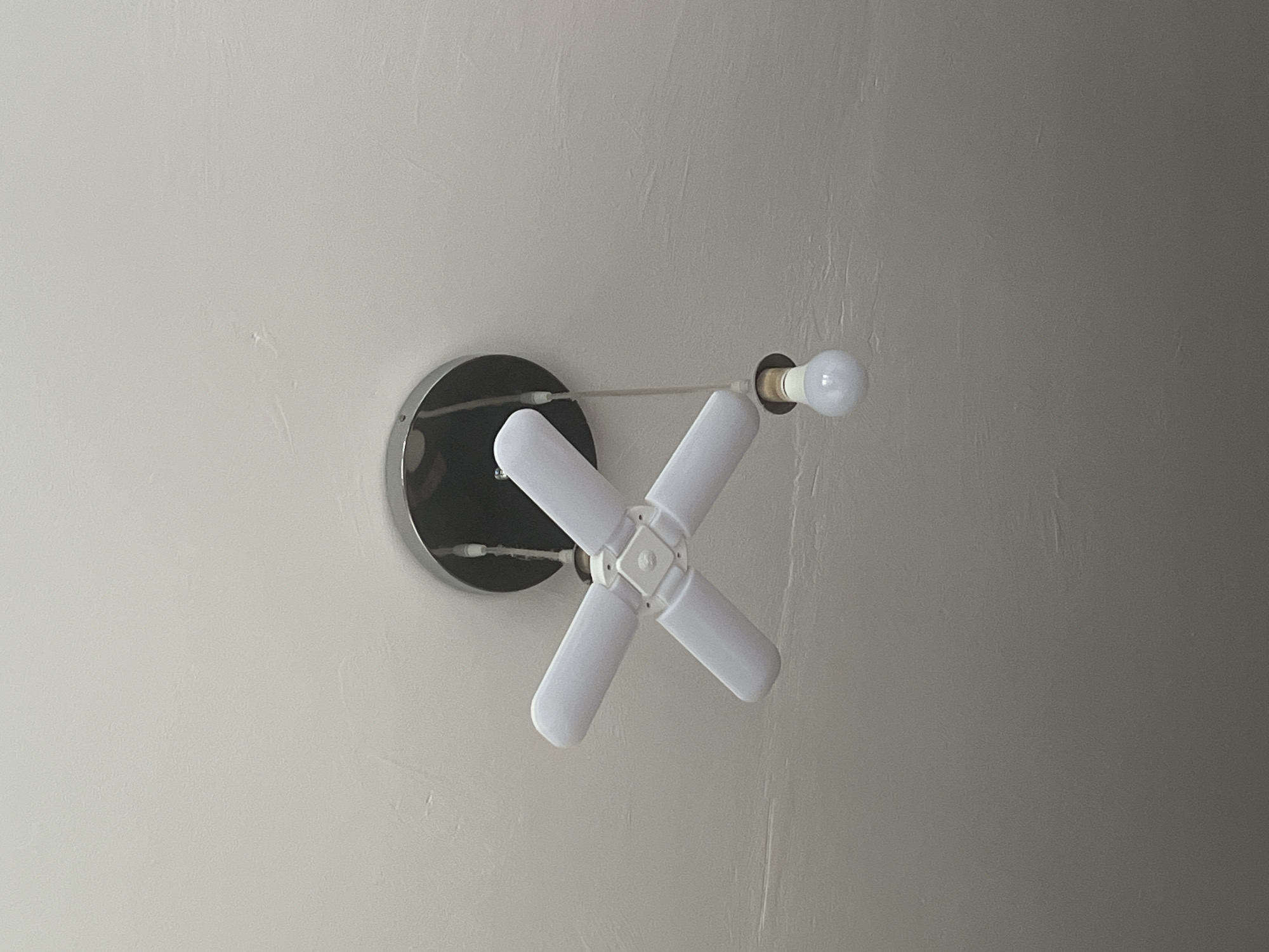 A white ceiling fan light fixture with four adjustable blades is mounted on a textured, off-white ceiling. A separate, dangling light bulb is suspended below the fixture by a thin cord. The scene is indoors, in an unspecified location.
